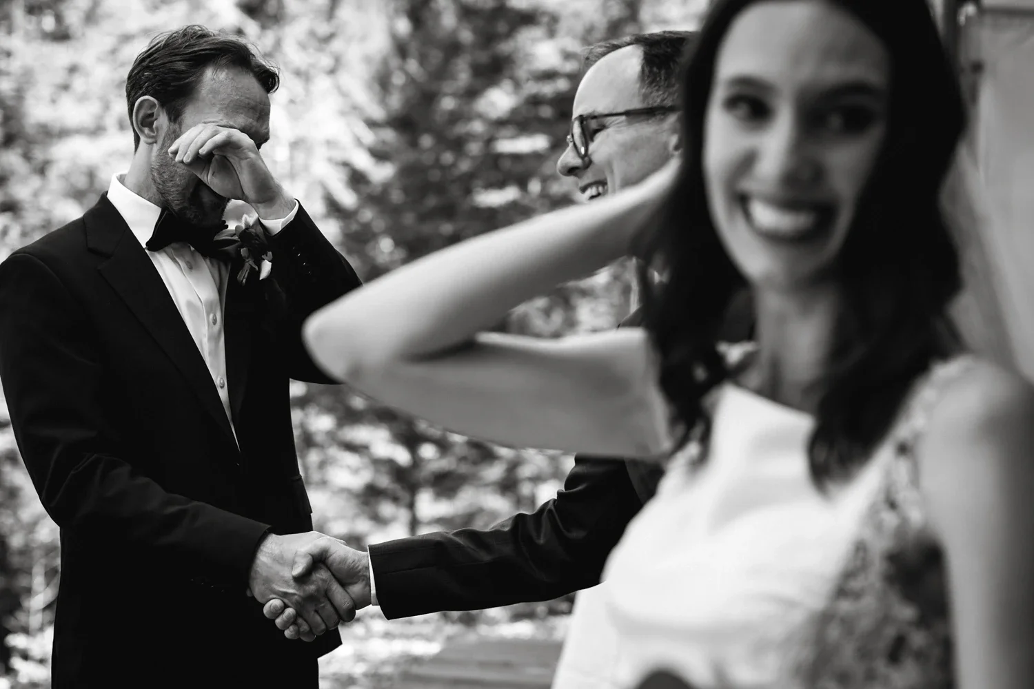 A groom in a tuxedo appears emotional, with his hand covering part of his face, as he shakes hands with another man. A woman in a wedding dress and veil is smiling in the foreground, out of focus, with trees in the background.