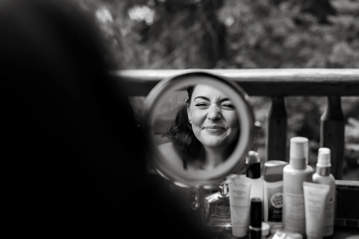 A woman making a funny face in a small round mirror with various bottles and skincare products on a table in front of her.