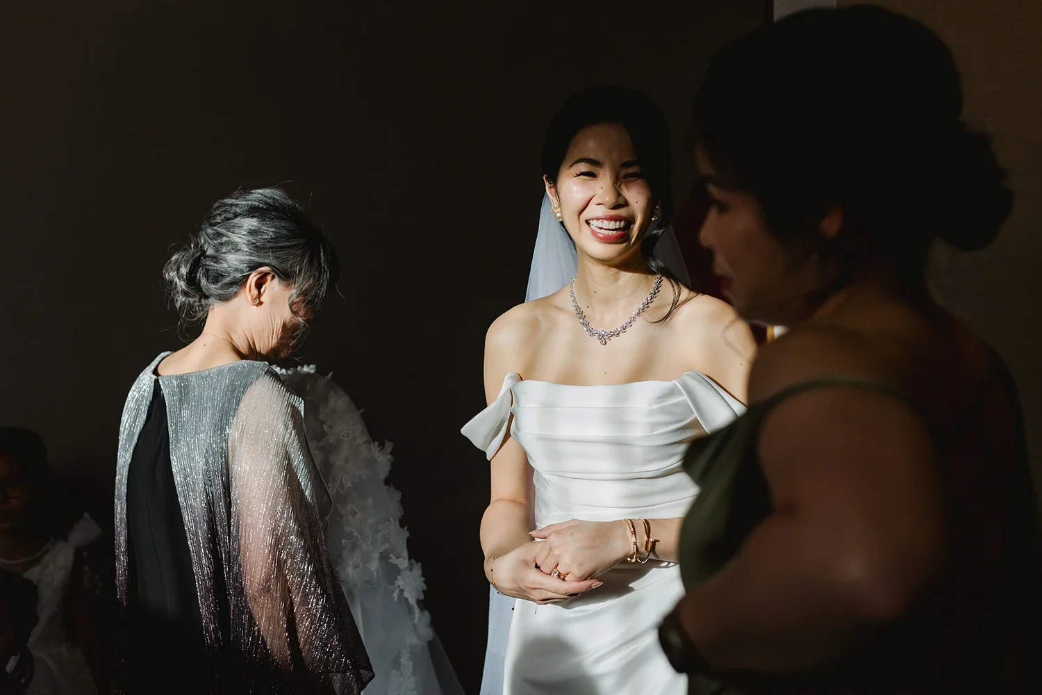 Three women, including a bride in a white wedding dress, are smiling and talking at a wedding reception.