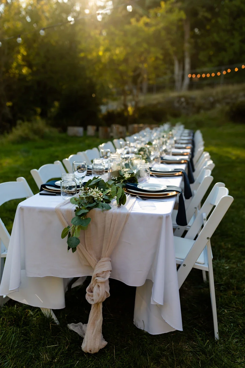 Elegant outdoor dining setup with a long table decorated with greenery and white flowers, set with plates, glasses, and black napkins, under string lights at sunset.
