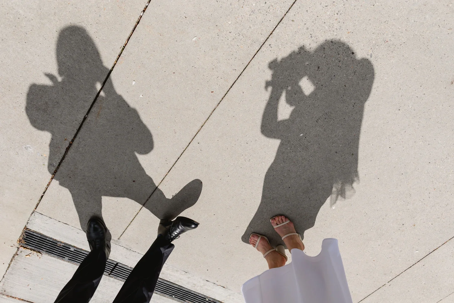 Shadows of two people, one with a camera and the other with a white dress, cast on a concrete sidewalk. The person on the left is wearing black shoes and pants, and the person on the right is wearing sandals and a white dress.