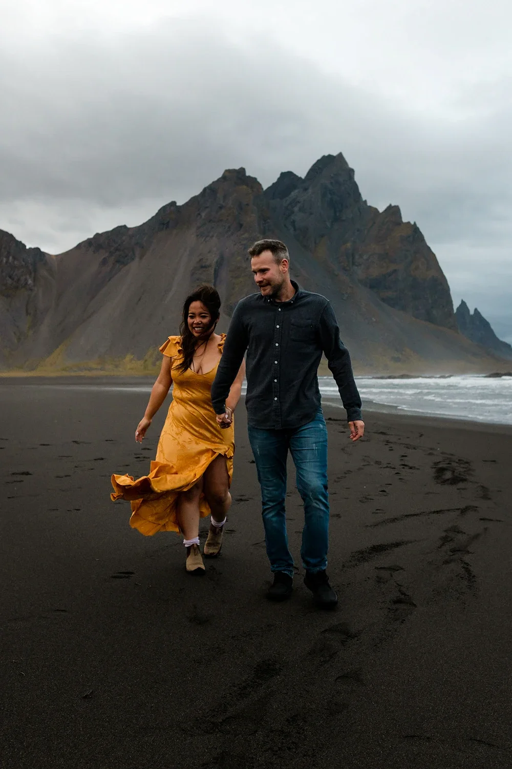 A couple walking hand in hand on a black sand beach with mountains in the background, overcast sky, woman wearing a yellow dress and the man in a dark shirt and jeans.