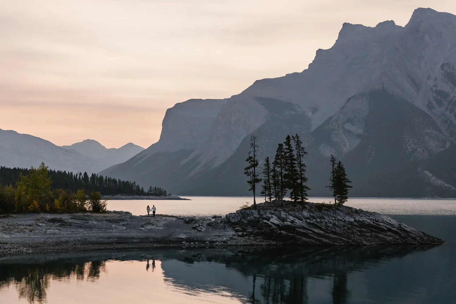 A serene lake surrounded by mountains at dusk, with a small island of trees and a person holding hands with a child on the shore.