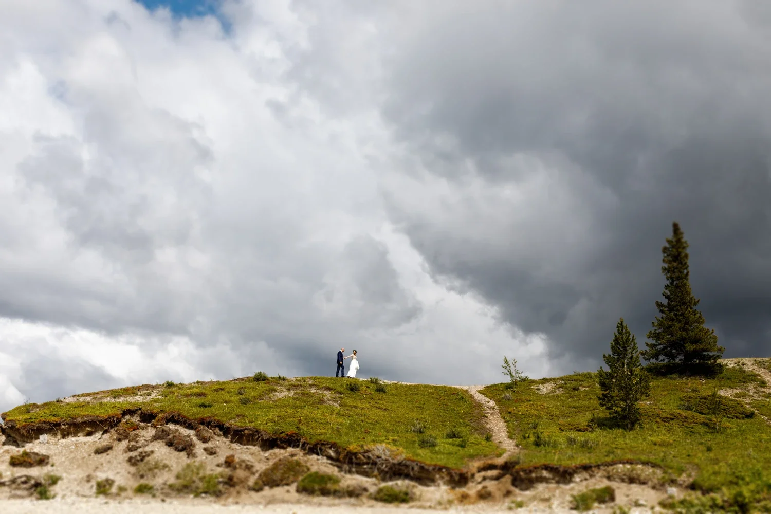 A bride and groom holding hands on a grassy hill, with a dramatic cloudy sky overhead and a few scattered trees.
