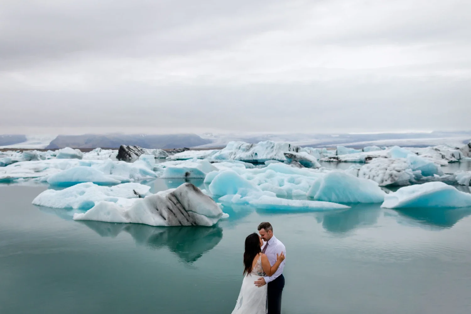A couple dressed in wedding attire standing close and embracing in a body of water surrounded by floating icebergs with a cloudy sky overhead.