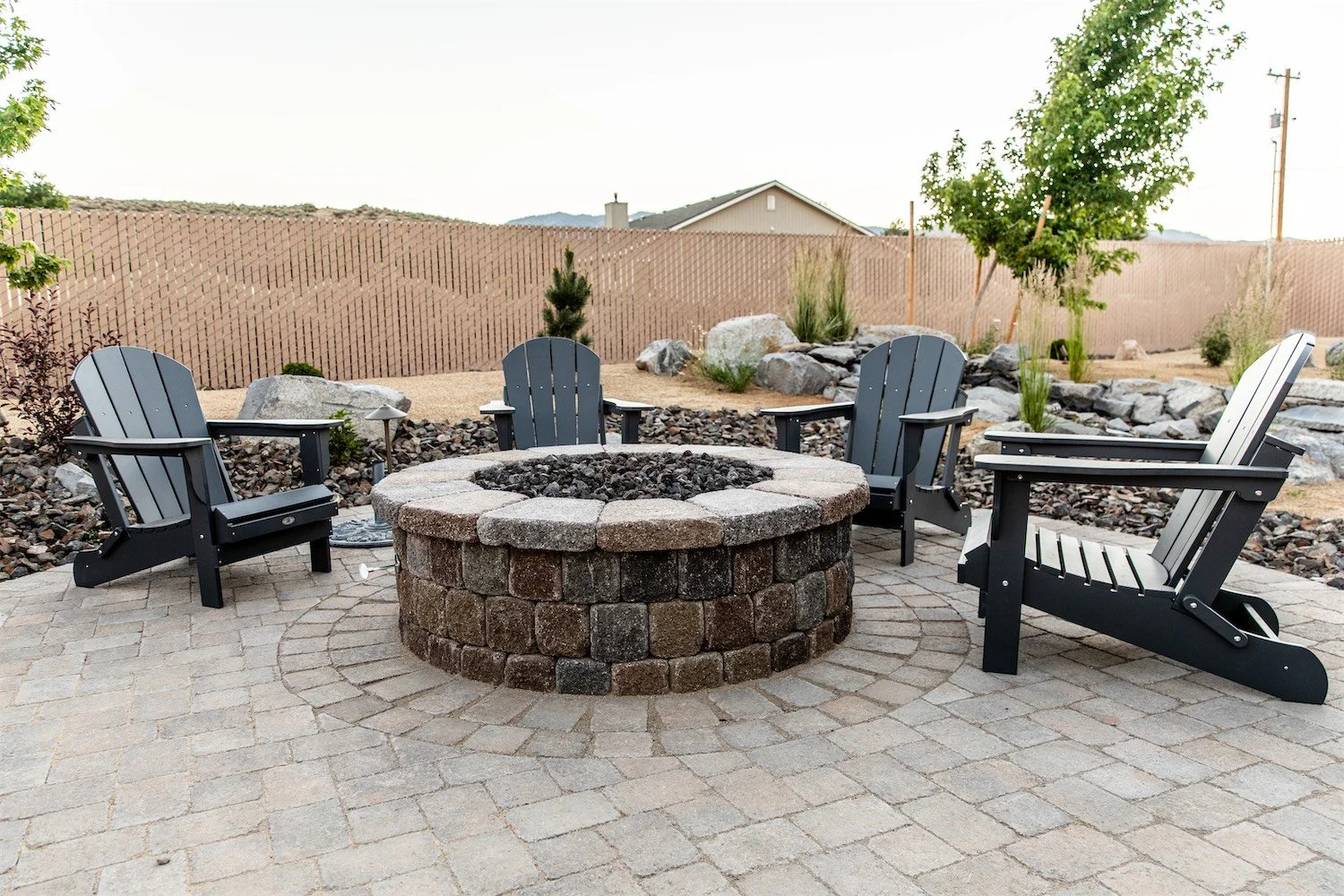 A backyard patio with four black Adirondack chairs surrounding a stone fire pit on a paved surface, with a landscaped background including rocks, plants, and a wooden privacy fence.