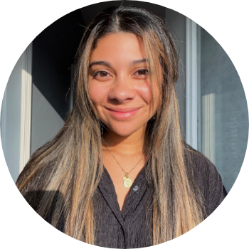 A young woman with long, light brown hair, smiling, standing near a window with sunlight illuminating her face.