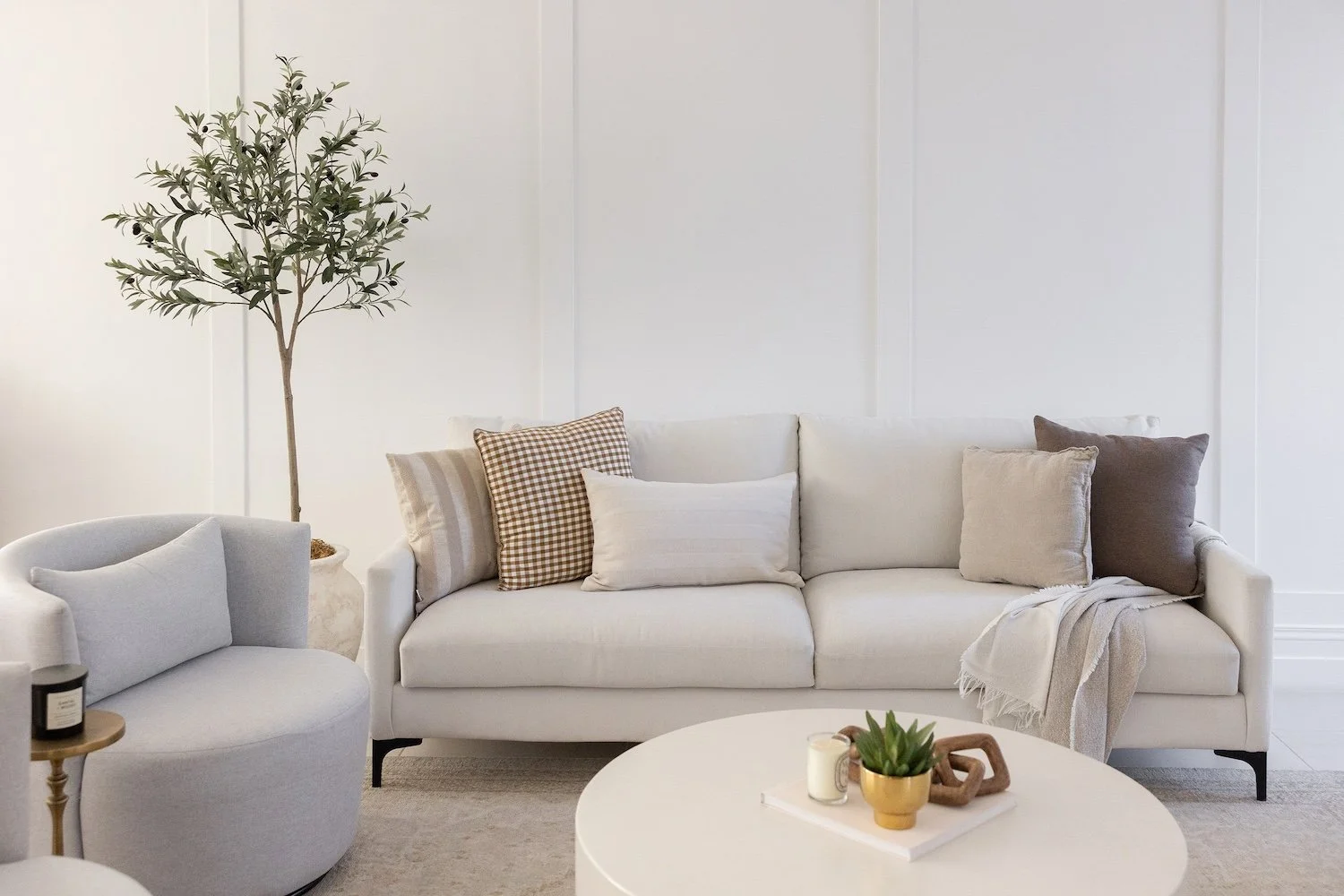 Minimalist living room with a white sofa, decorative pillows, a round coffee table with a plant and candles, a white armchair, and an indoor potted tree against a white wall.