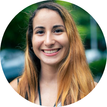 A young woman with long, straight, light brown hair smiling outdoors with a blurred green background.