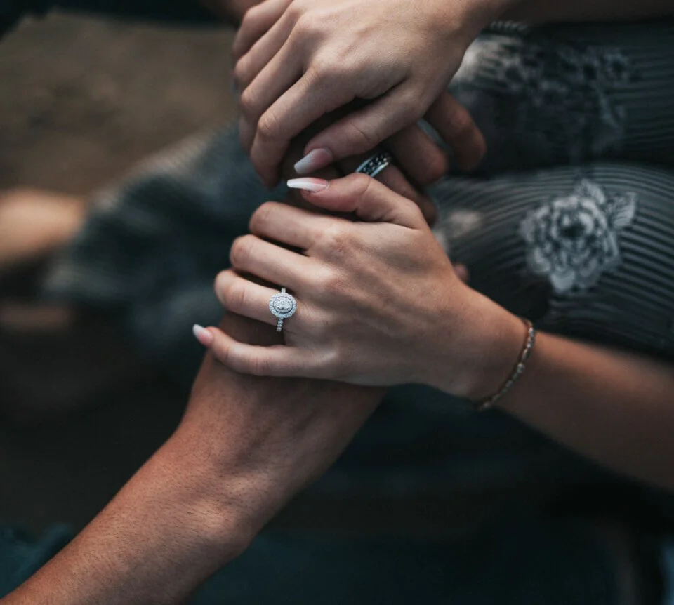 Close-up of two hands, one with a large engagement ring, gently holding the other hand, indicating a romantic or engagement moment.