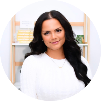 A woman with long dark hair and a white top standing in front of a bookshelf with yellow and white books.