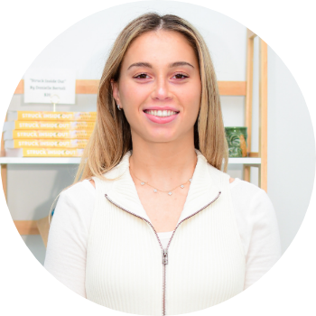 A young woman with long blonde hair smiling in a bright indoor setting, possibly a store or cafe, with shelves and books in the background.