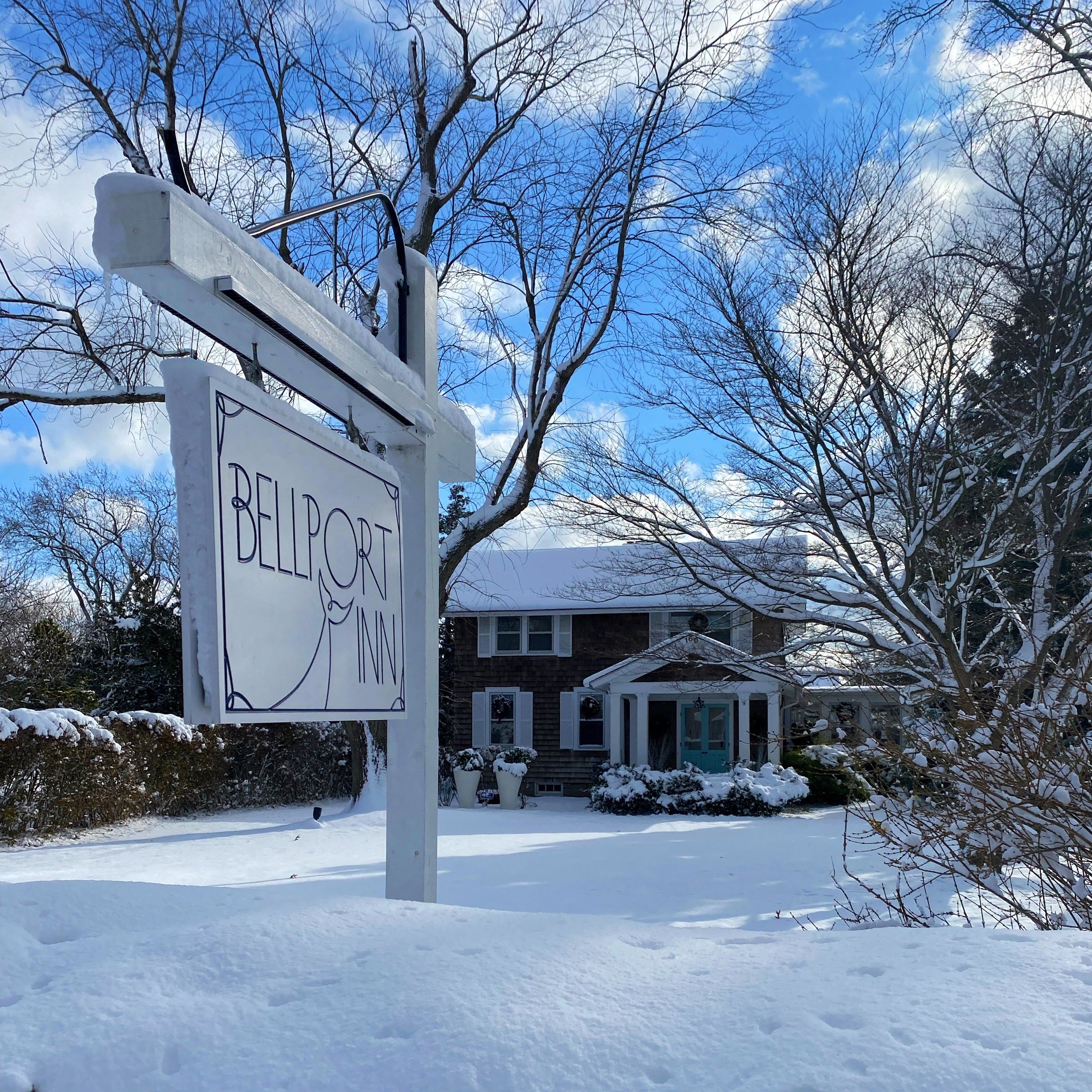 A snow-covered yard with a sign reading 'Bellport Inn' hanging from a white post, with a house in the background and snow-laden trees surrounded by a blue sky with clouds.