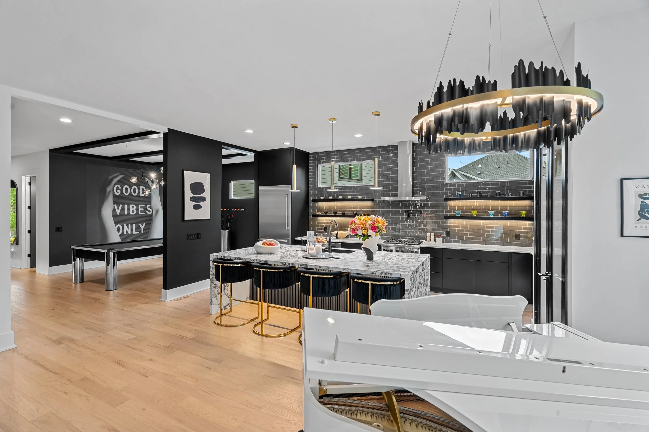 Modern kitchen with white marble island, black cabinets, gray subway tile backsplash, black and gold bar stools, vibrant flower arrangement, black chandelier, and light wood flooring.