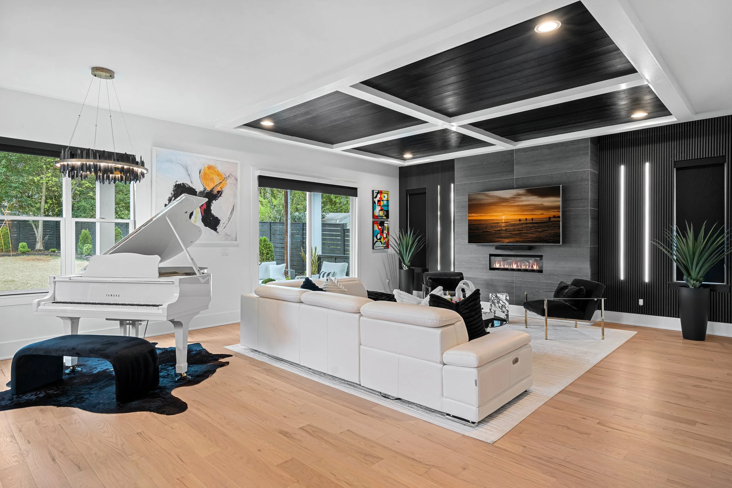 Modern living room with white piano, white sofas, black and white chairs, wooden floor, black ceiling accents, large windows, TV with sunset on screen, abstract wall art, indoor plants, and contemporary decor.