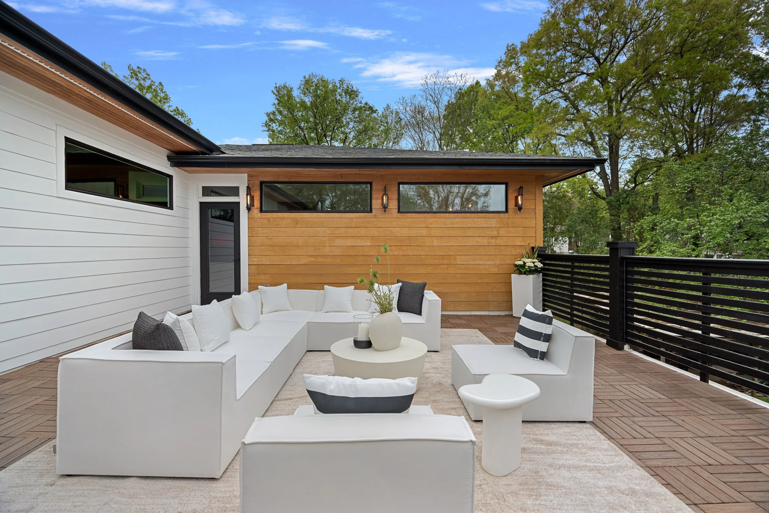 Modern outdoor patio with white sectional sofa, black and white patterned cushions, round coffee table with candles, potted plant, surrounded by black fence and wooden wall, with trees and blue sky in background.