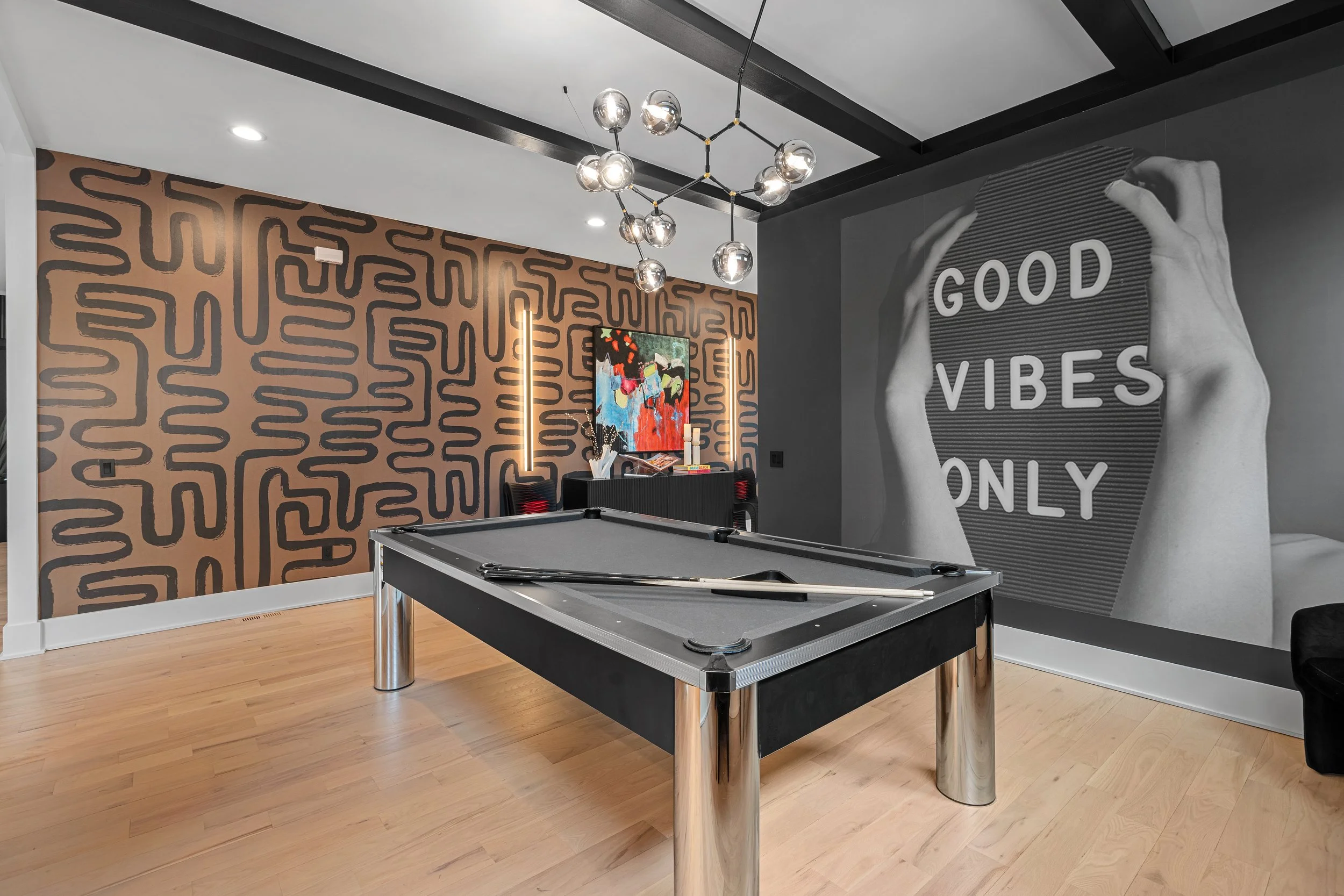 Interior of a modern living room featuring a gray pool table, a wall with black and tan abstract pattern, and a large mural with a hand holding a textured surface with the words 'Good Vibes Only'. There is a minimalist chandelier with multiple glass 