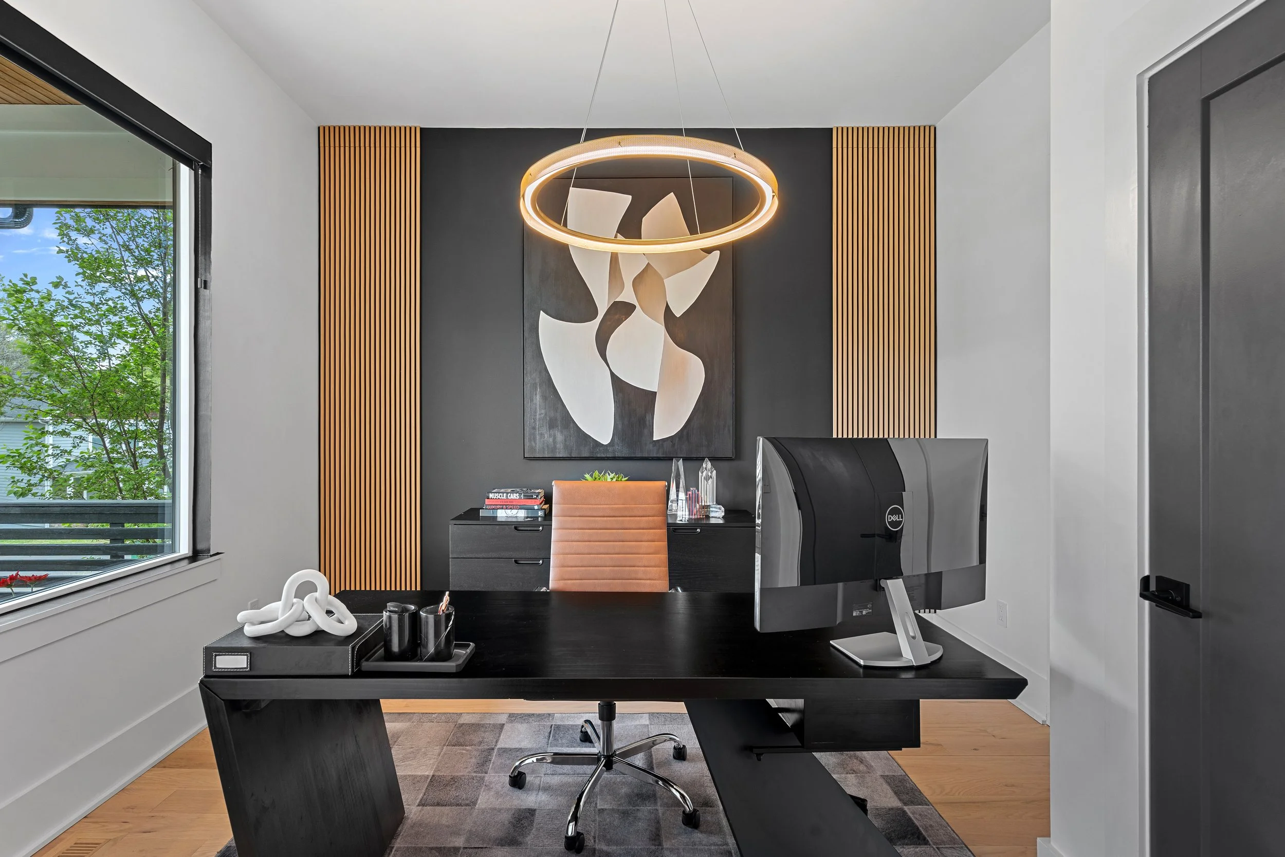 Modern office with black desk, orange chair, computer monitor, decorative items, abstract artwork on dark wall, wooden wall panels, and large window showing trees outside.