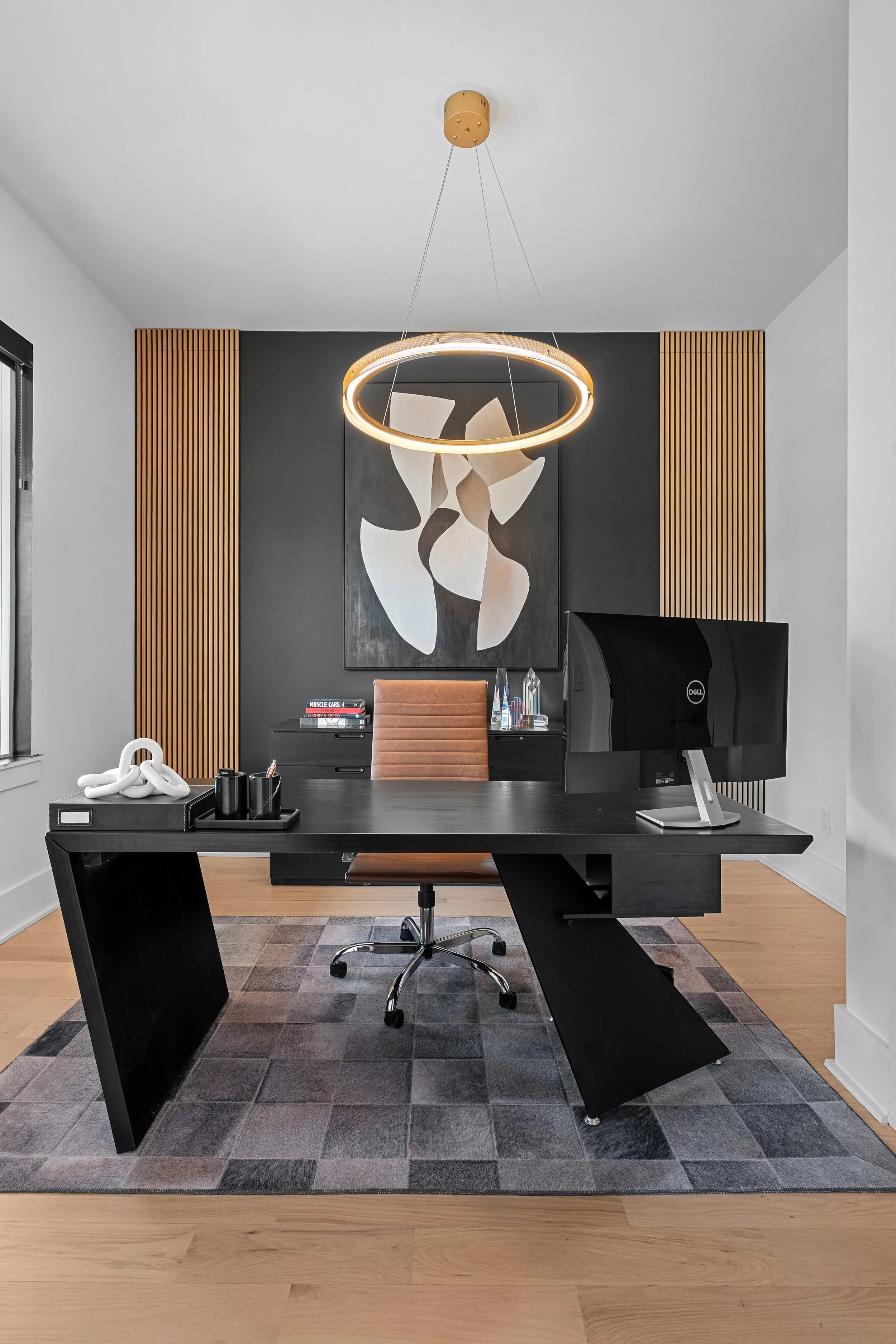 Modern office workspace with a black desk, a chair, computer monitor, decorative objects, and wall art, illuminated by a contemporary hanging light fixture.