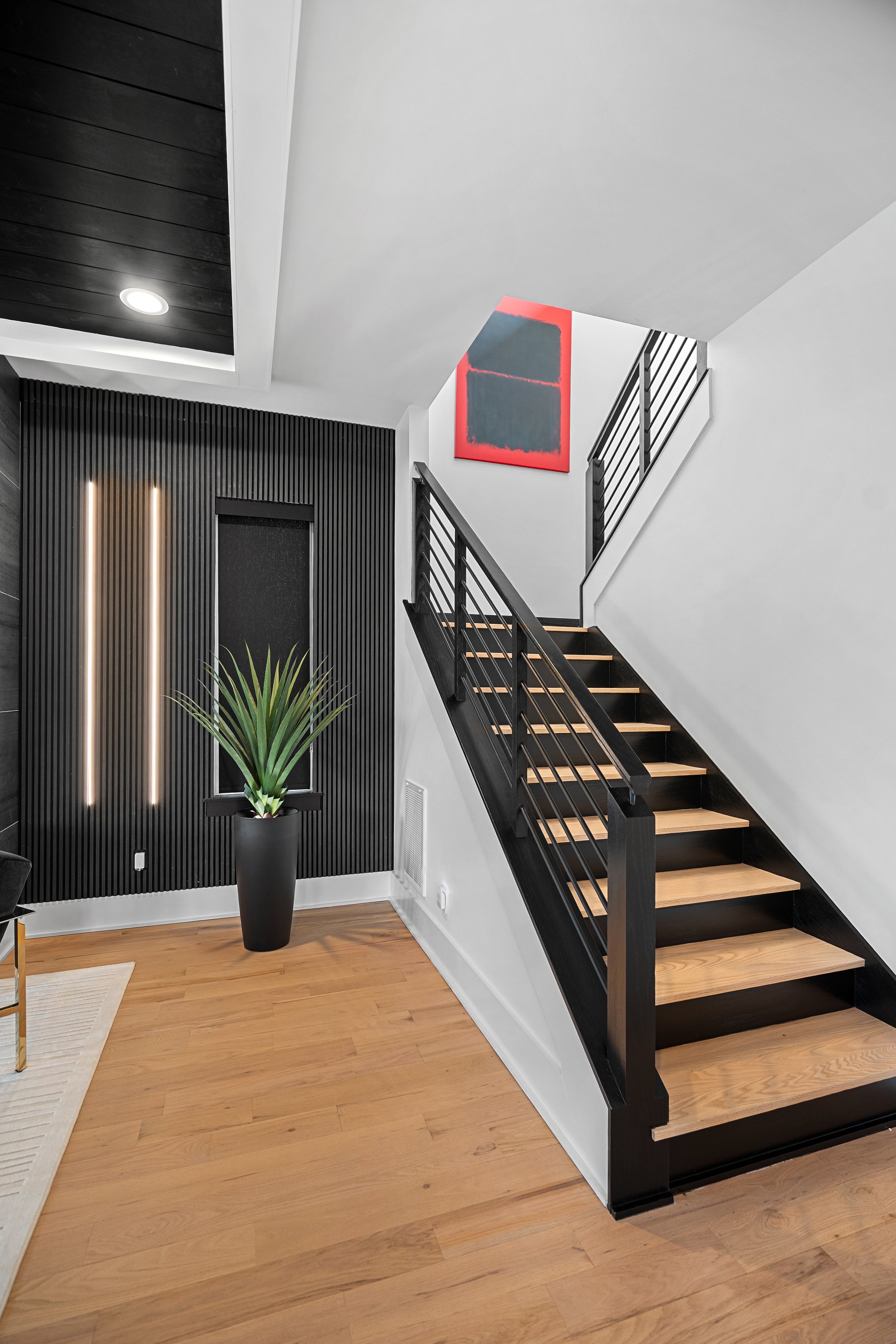 Interior staircase with black railing and light wooden steps, black wall with vertical slats, a potted plant, and abstract artwork hanging on the wall, with hardwood flooring.