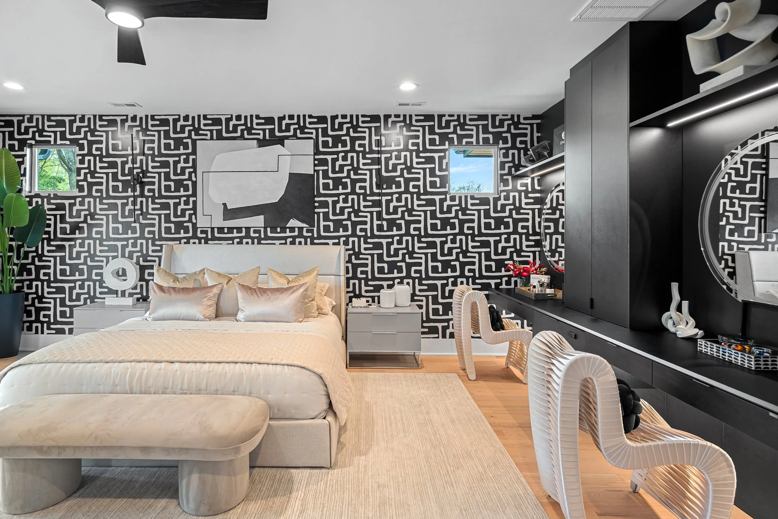 Modern bedroom with black and white geometric patterned accent wall, white bed with beige pillows, white bedside table, large green plant, artwork, black storage unit, and decorative white chairs.