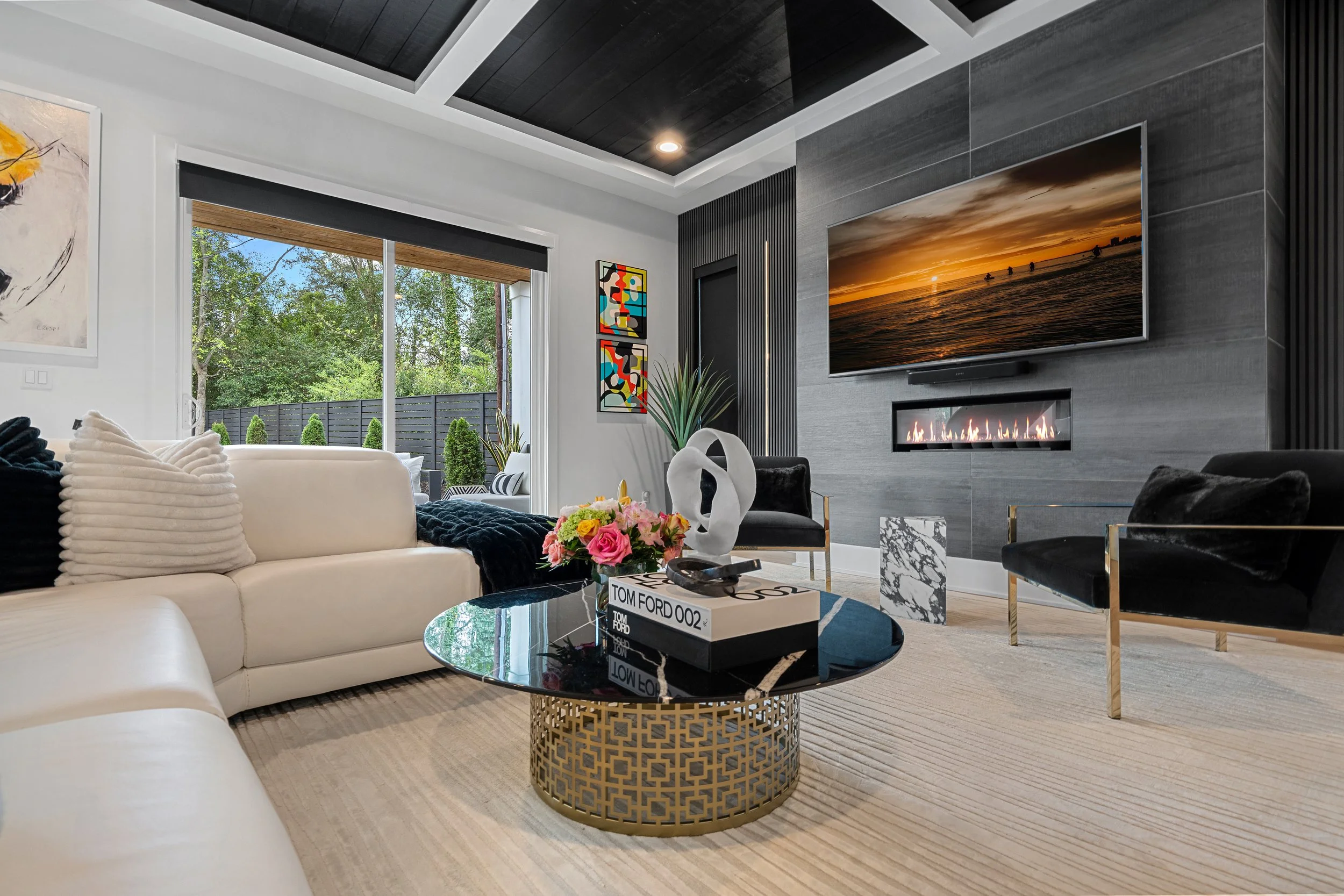 Modern living room with white sofa, black velvet chairs, abstract art, large window with outdoor greenery, wall-mounted TV displaying a sunset, fireplace, decorative plants, and coffee table with a flower bouquet and books.
