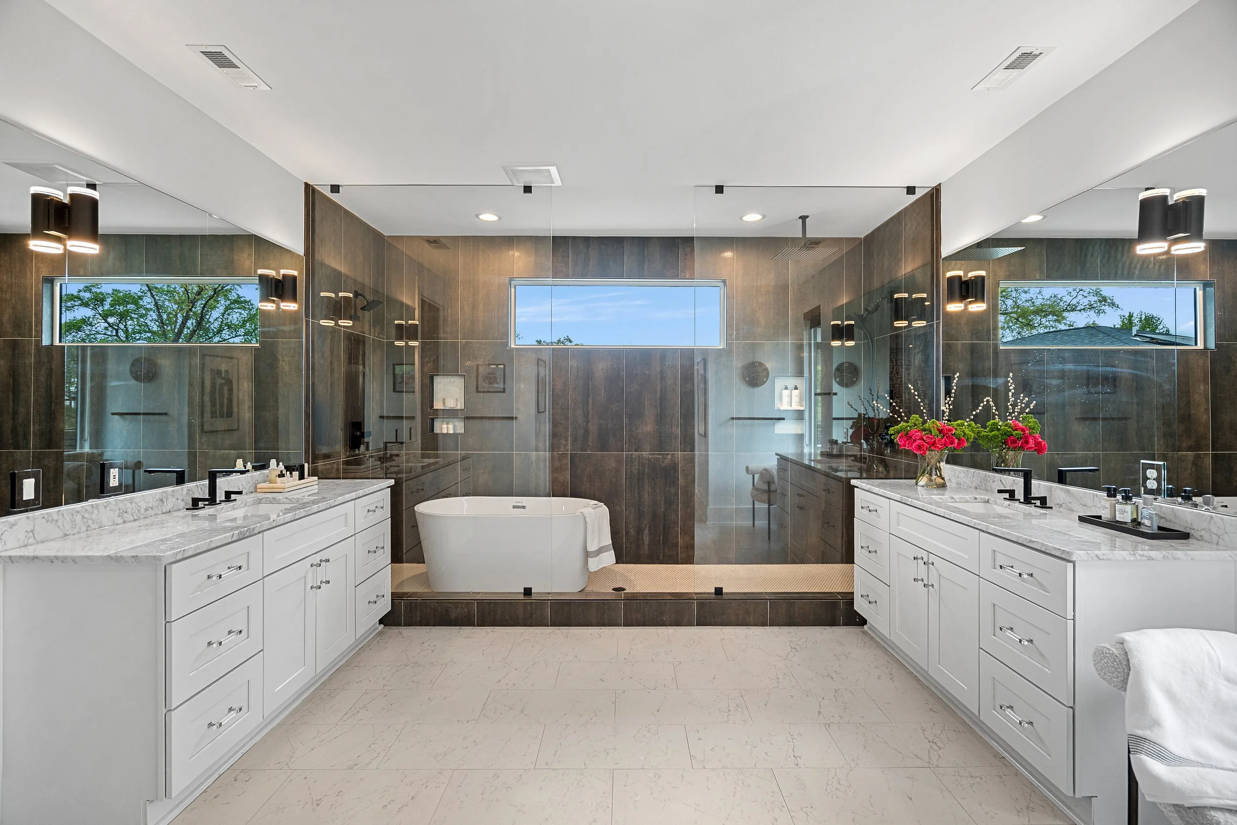 A modern bathroom with a large mirror, white double vanity with marble countertop, black faucets, and a bathtub in the center. The bathroom has dark tiled walls, two long windows, and decorative flowers on the right counter.