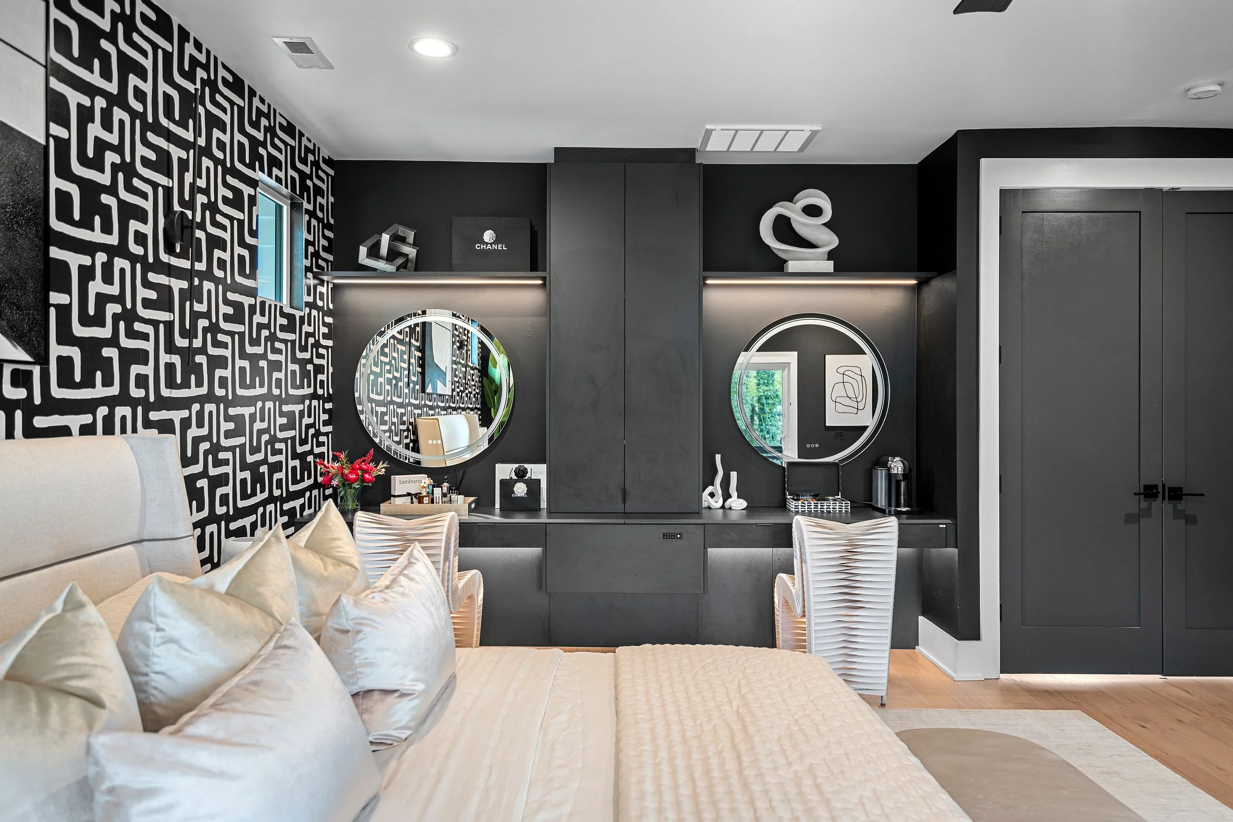 A modern interior with black and white decor, includes a beige bed with many white pillows, a black wall with circular mirrors, decorative items, and art, and a patterned black and white wall on the left.