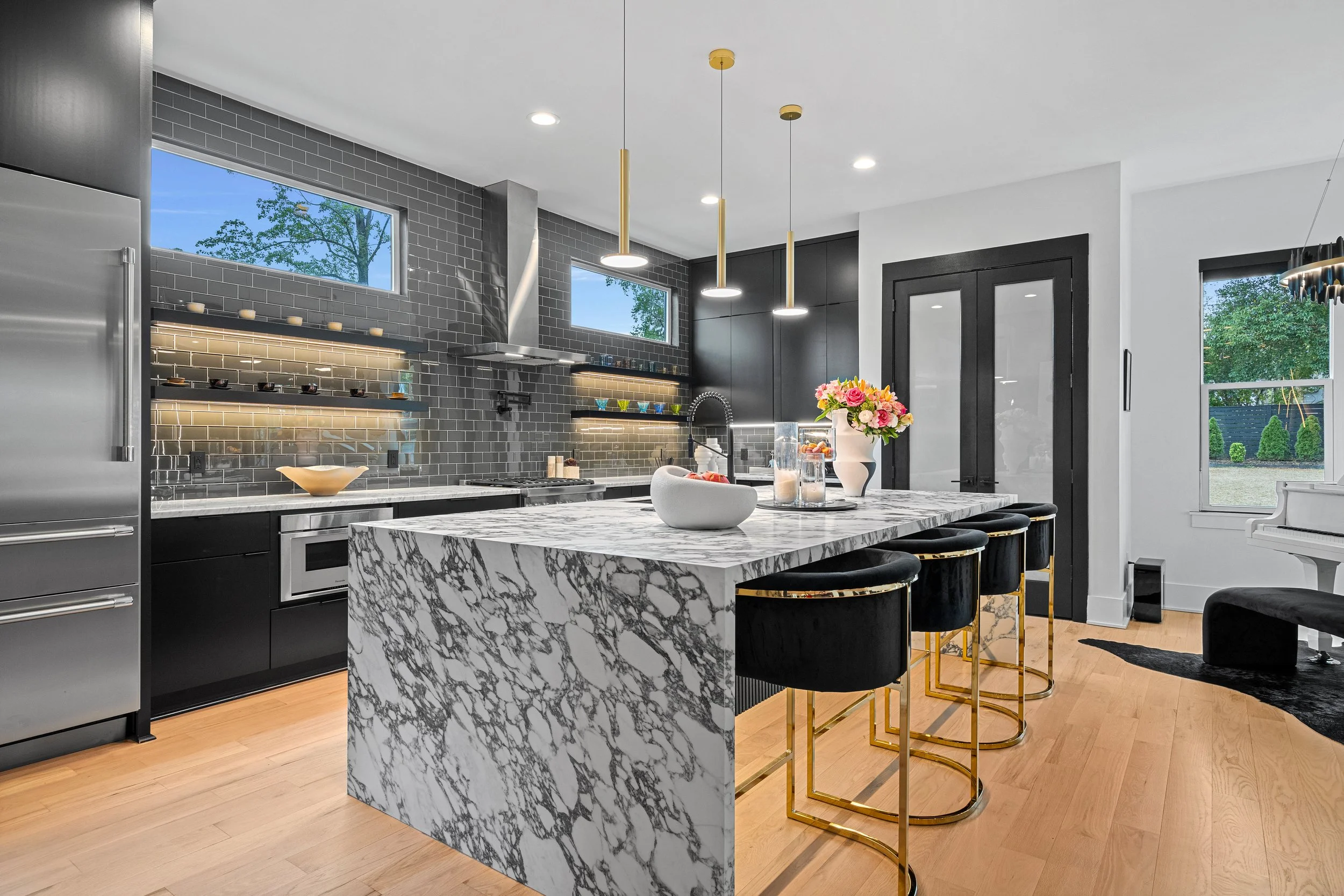 Modern kitchen with black and gray cabinets, marble island, black tile backsplash, and gold bar stools with black cushions.