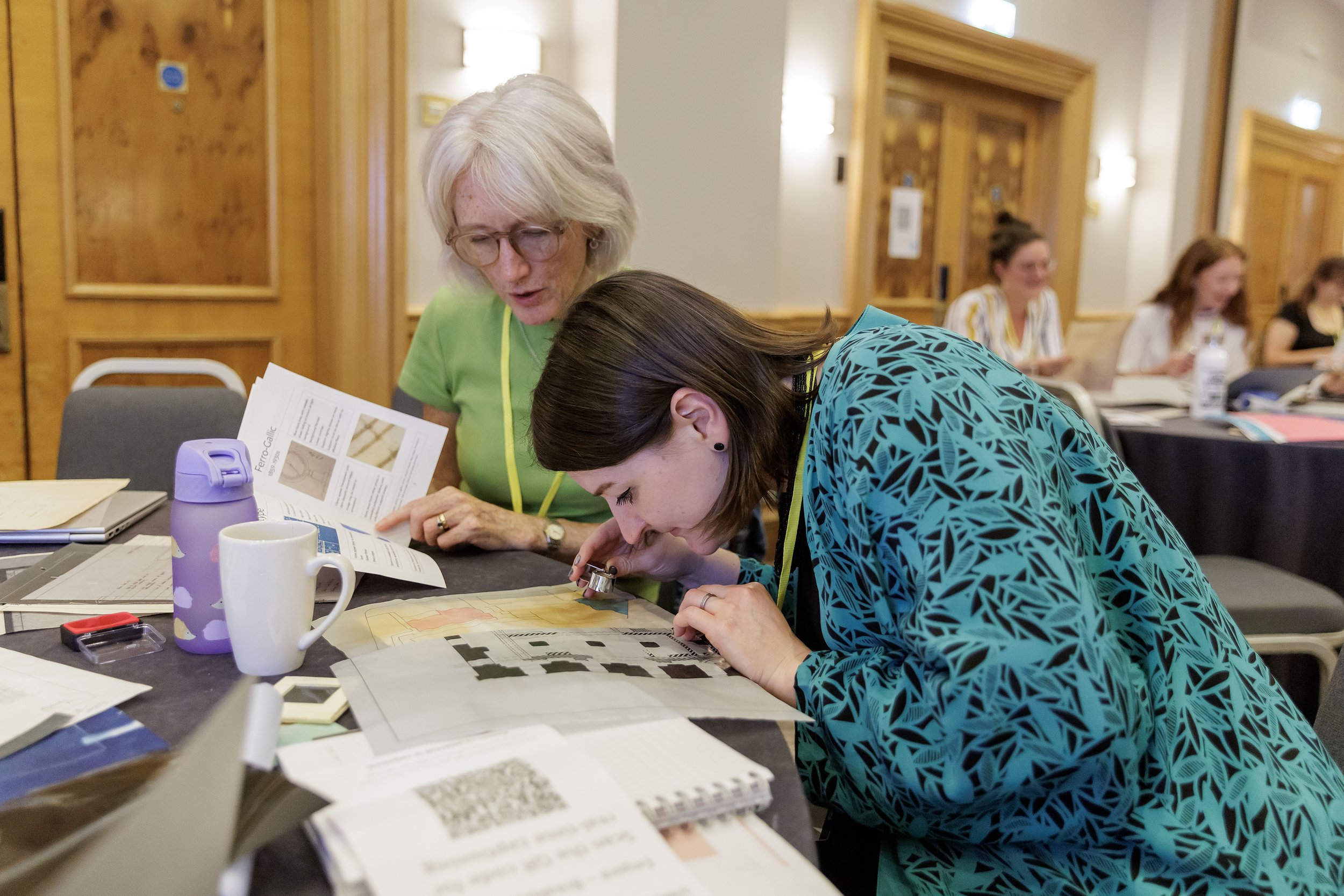 Image shows two women at a table covered in papers - one is holding an eyeglass to her eye to look closely at some plans - the other is pointing to some text in a small booklet