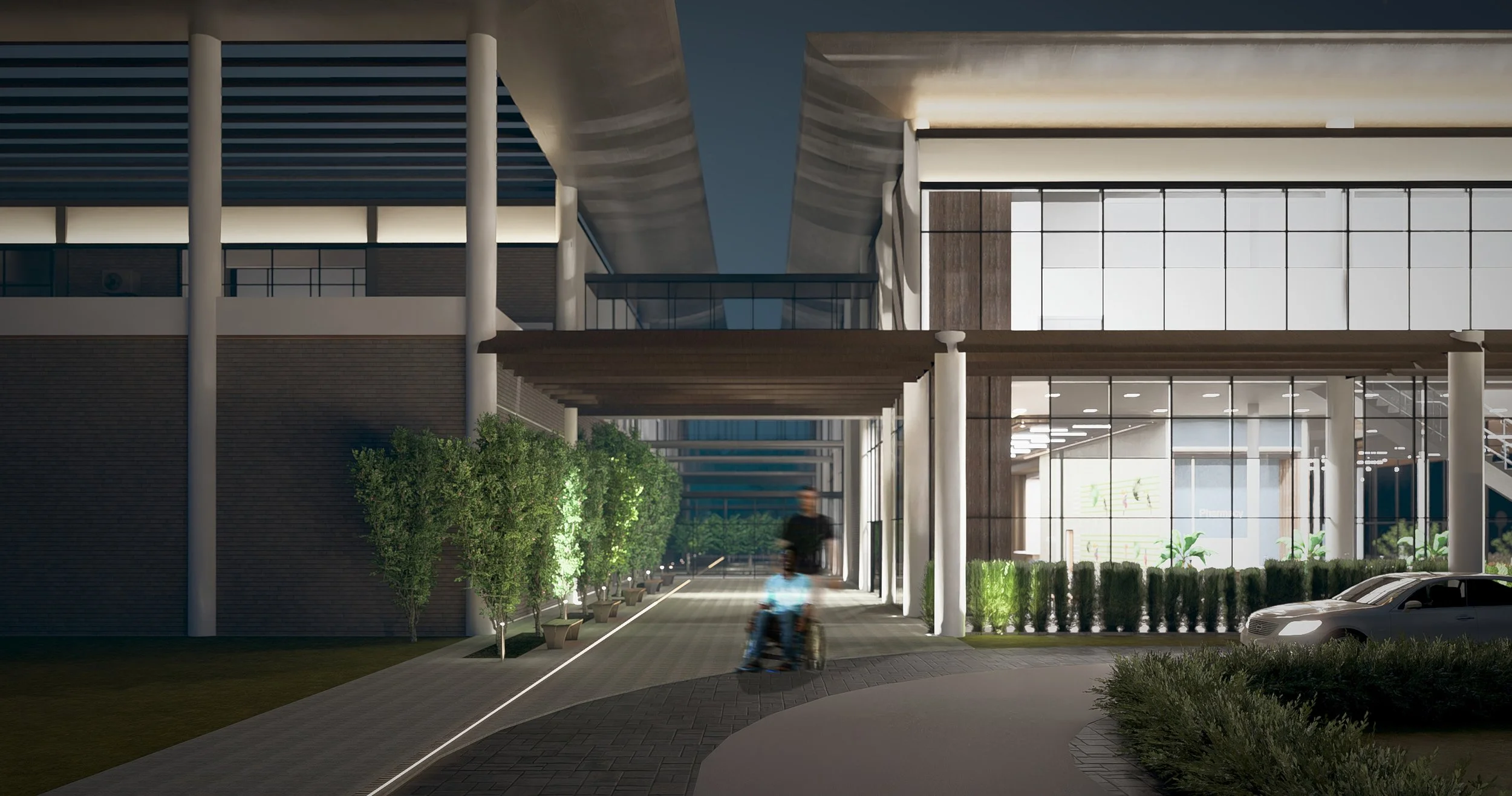 Nighttime view of a modern building with large glass windows, illuminated interior, and a sidewalk with trees and bushes; a man in a wheelchair is in motion along the sidewalk