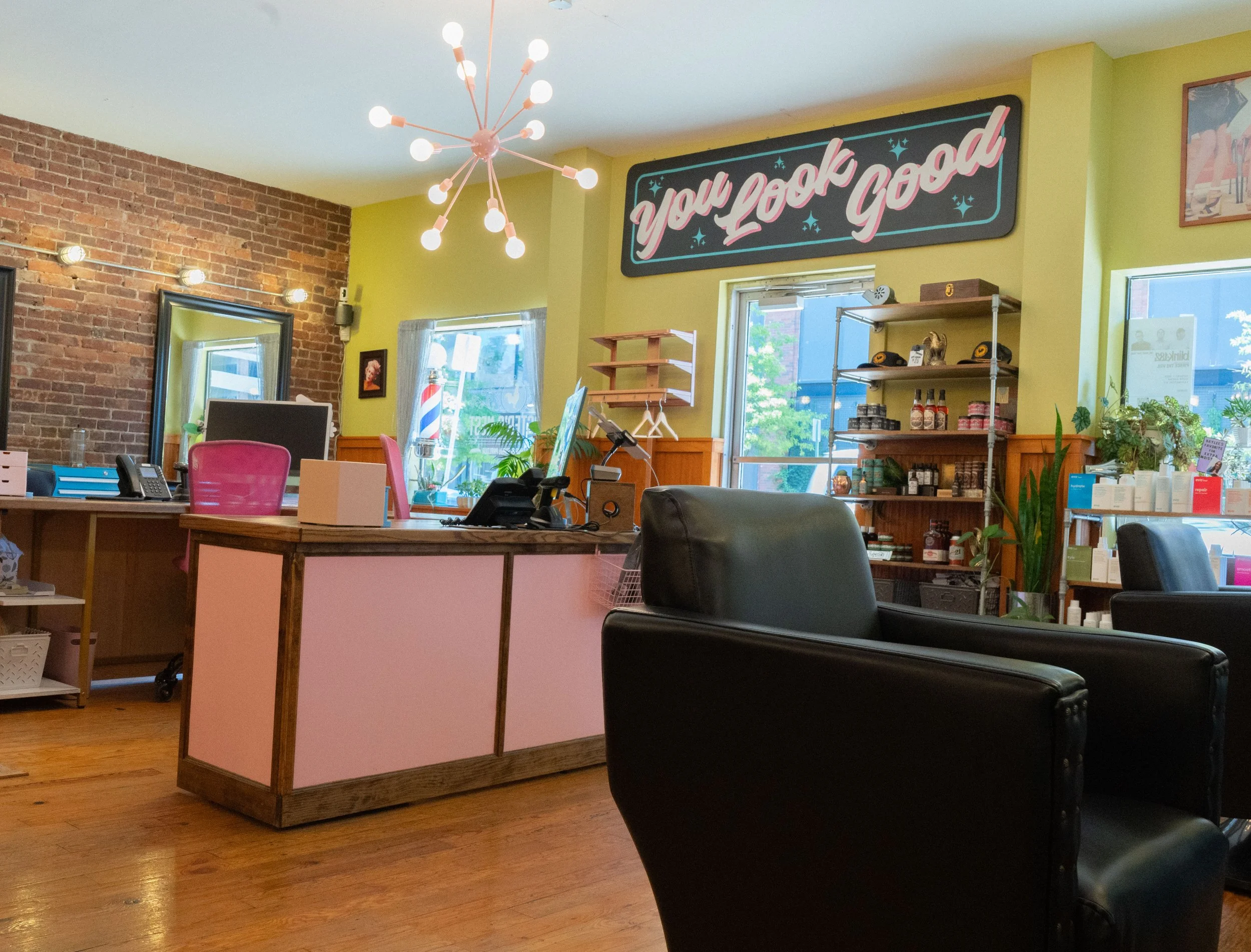 Interior of Lexington's best salon with a pink reception desk, black leather chairs, shelves with products, a neon sign that says "You Look Good," and a modern pink chandelier.