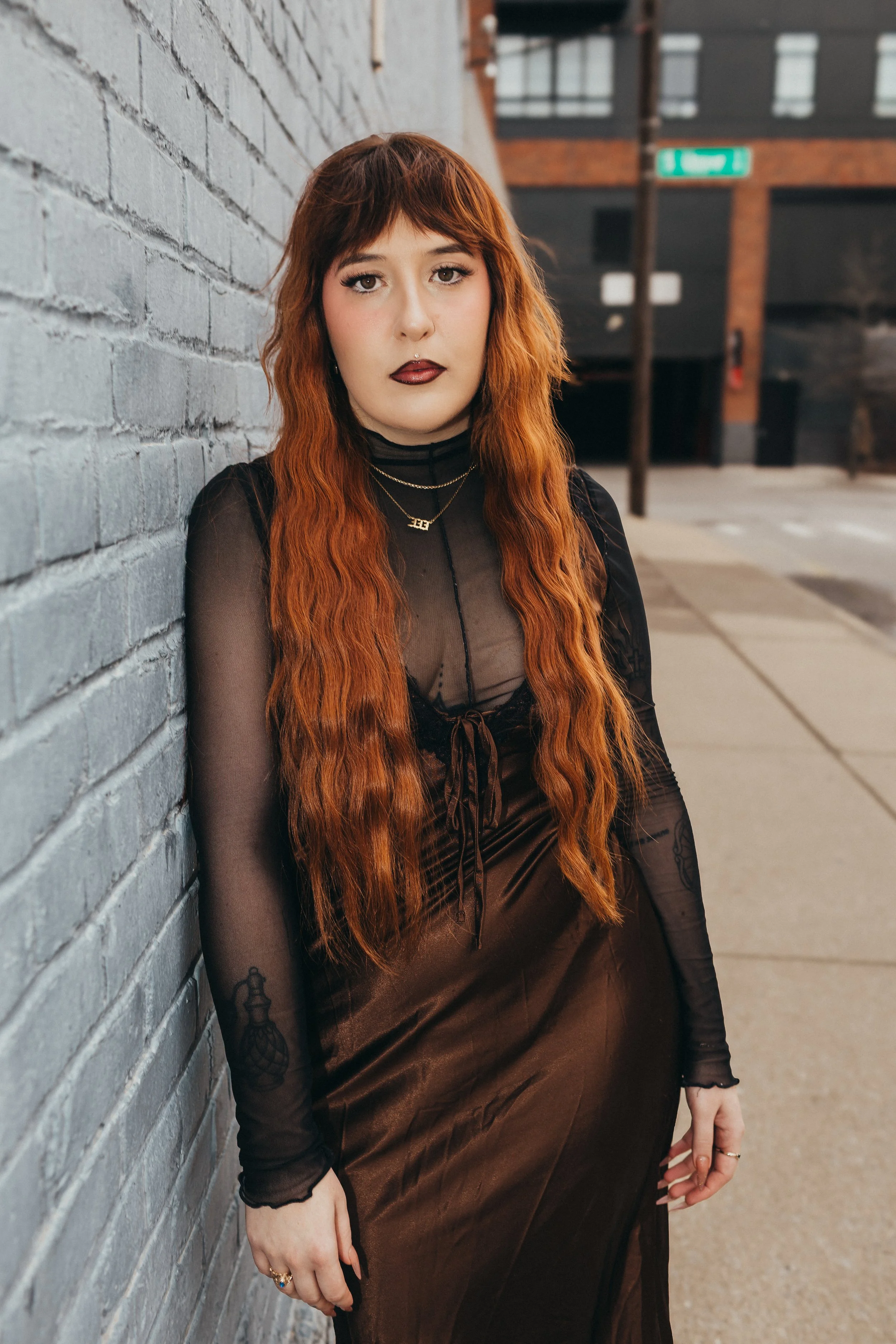 One of Lexington's best stylist is sitting on outdoor metal stairs, smiling, wearing a black tank top with a motorcycle graphic, gray cargo pants, and white sneakers, leaning against a gray brick wall.