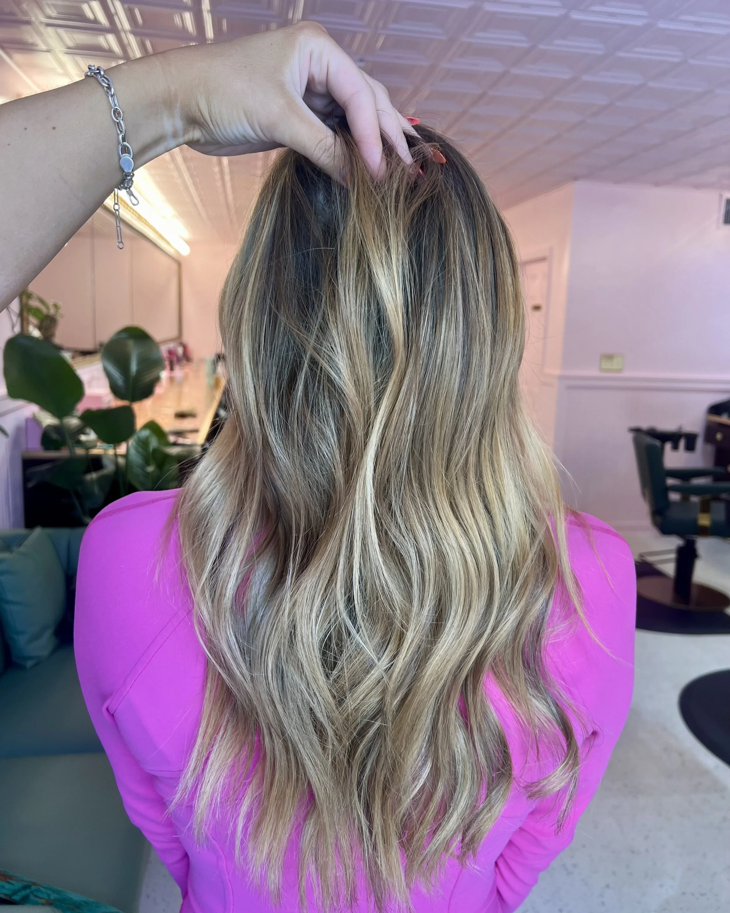 A woman with long, wavy blonde hair wearing a pink shirt, with her back turned. Someone is holding her hair at the top of her head. The background shows a room with plants, a couch, and chairs.