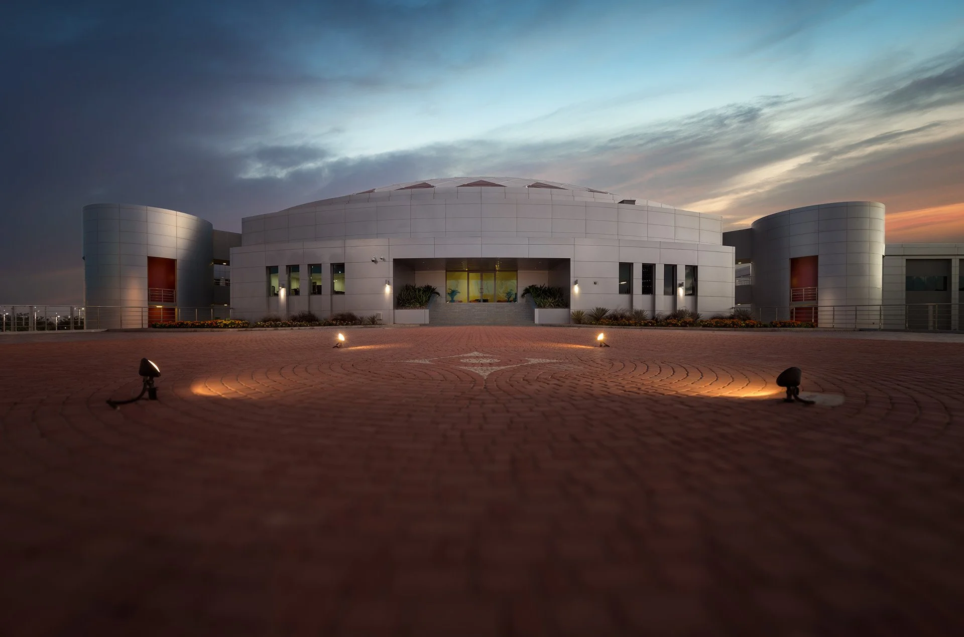 Symmetrical dusk exterior photograph of the Aspire Zone Ladies Sports Hall building showing illuminated paved forecourt, curved facade wings, and main entrance feature.