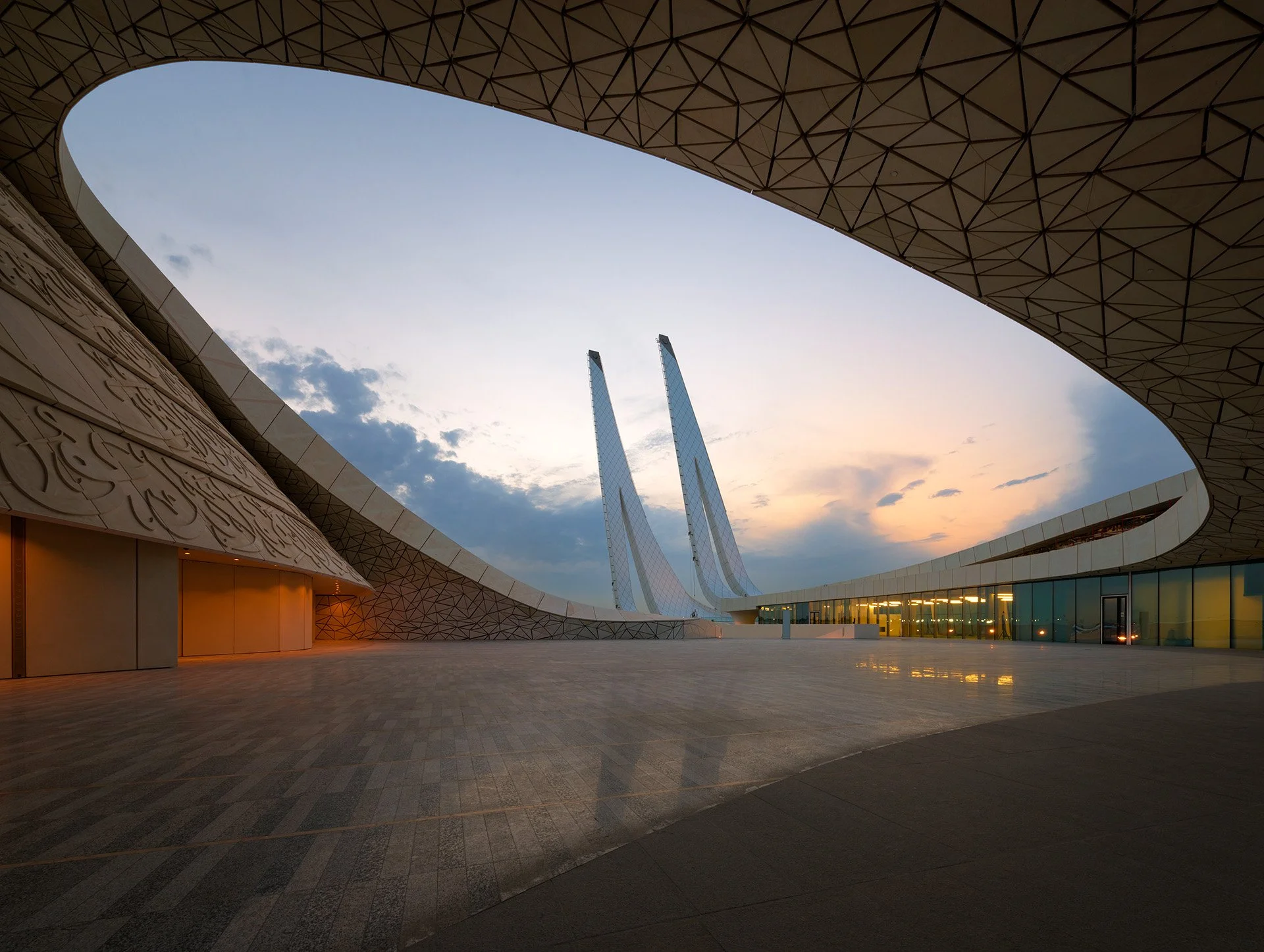 Wide angle twilight photograph of the Minaretein Center courtyard in Education City showing the calligraphic wall facade, geometric ceiling roof cutouts, and twin minarets.