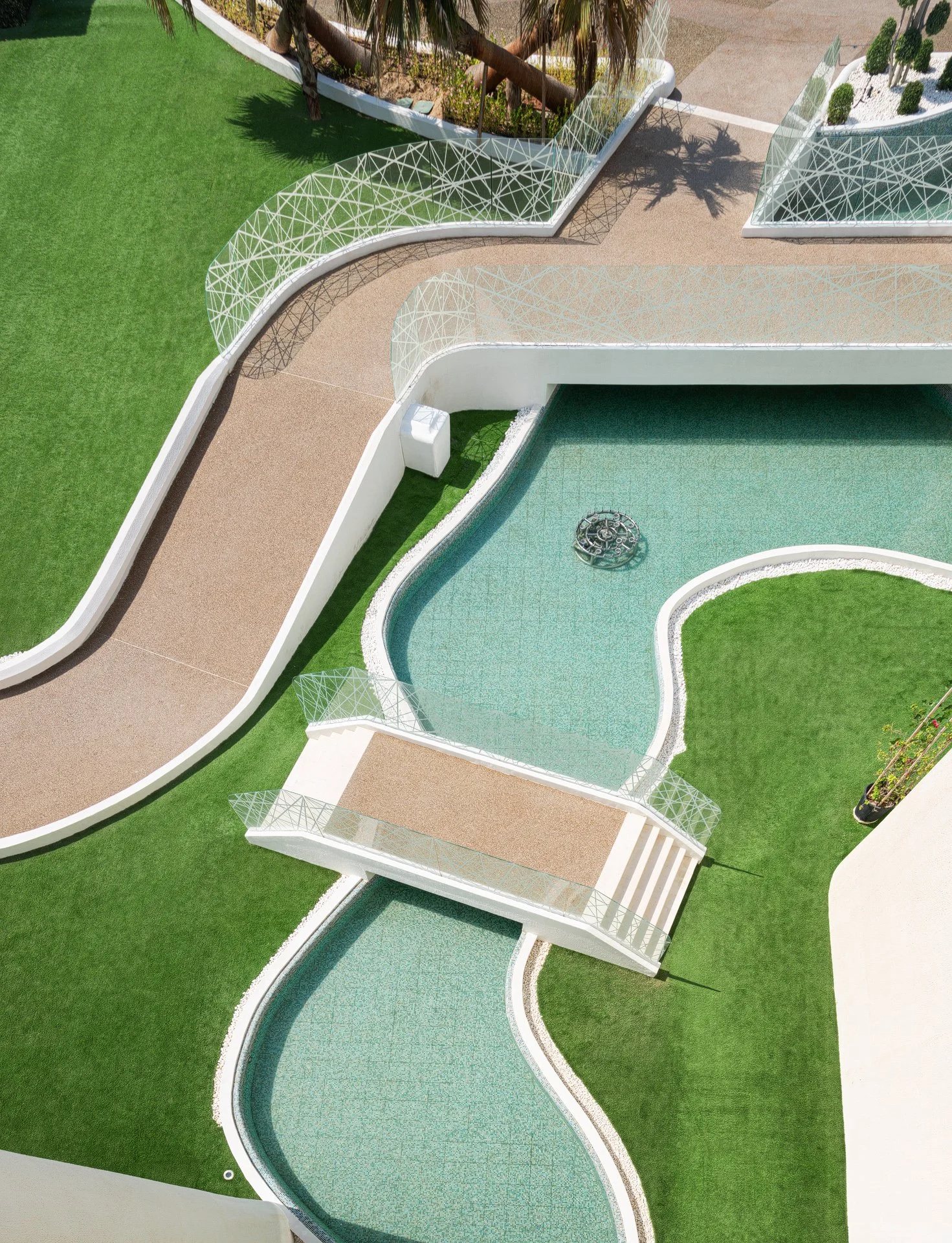 Drone-style top down architectural photo of meandering pool channels white pedestrian bridge and synthetic turf lawns by Hammad Iqbal