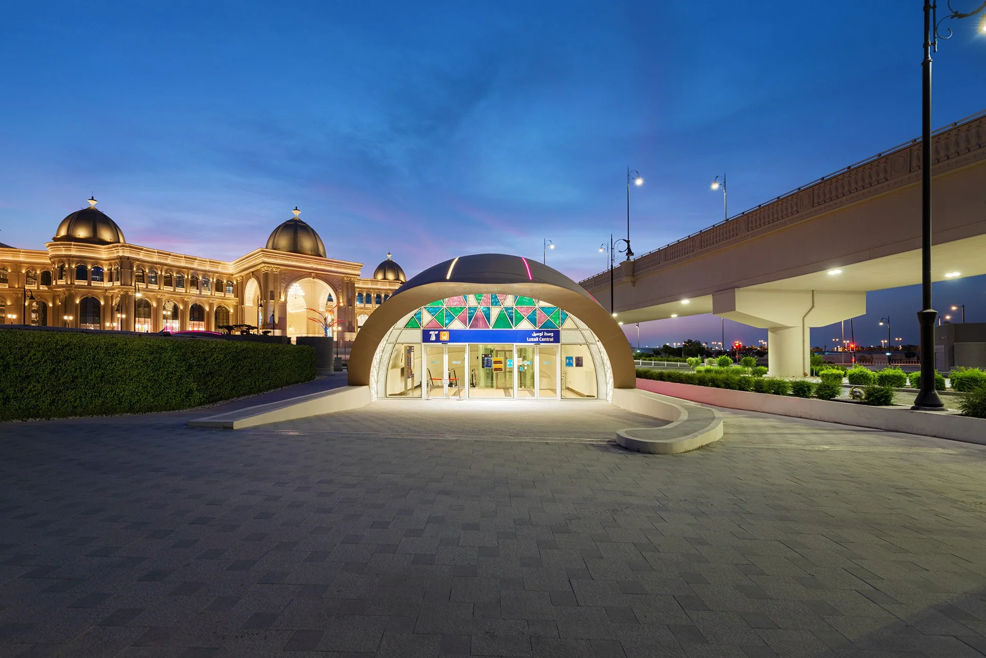 Head-on dusk architectural photograph of the domed Lusail Central LRT station entrance featuring colorful geometric glass paneling, set beside Place Vendôme Mall and an elevated roadway.