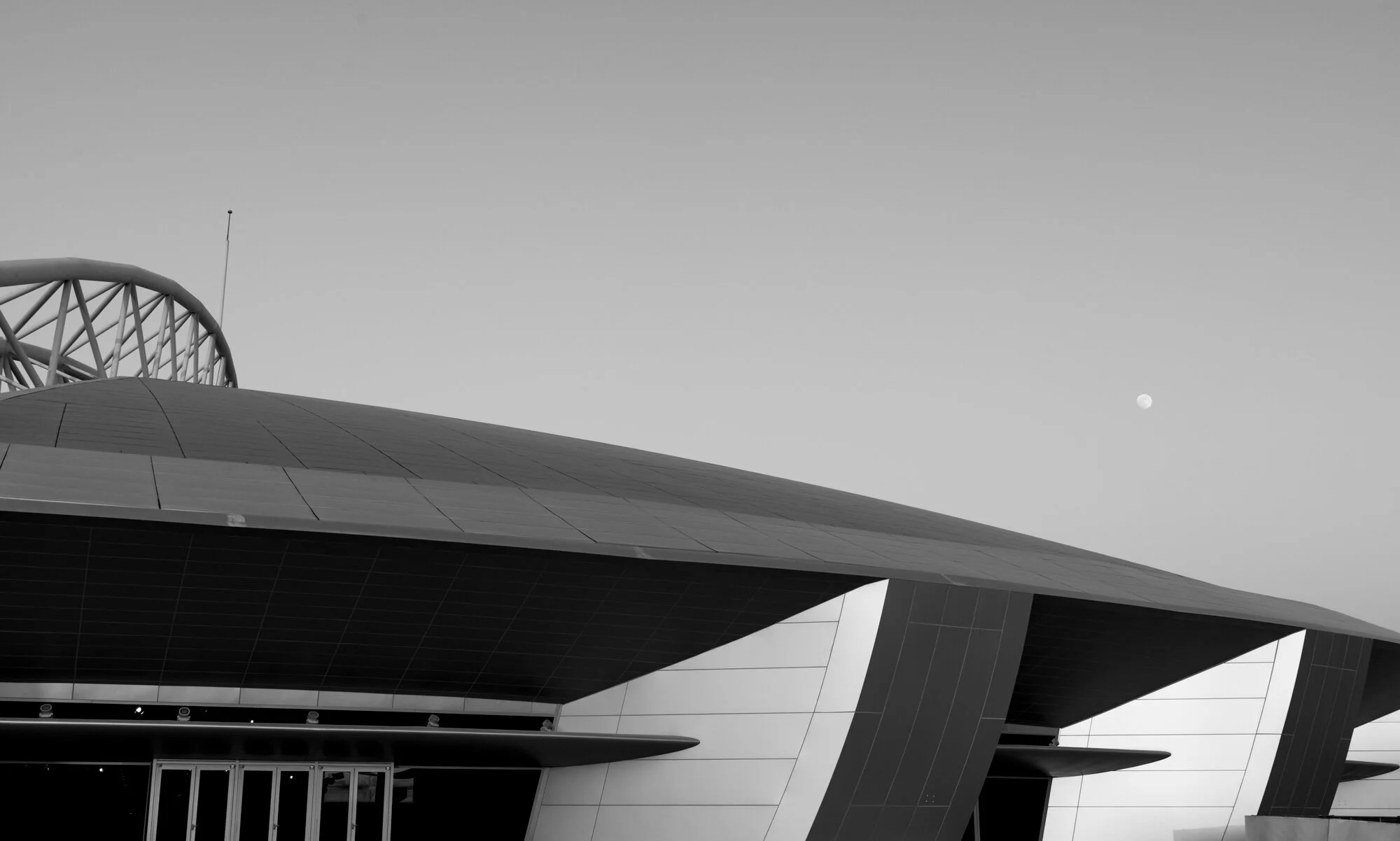 Fine art black and white shot of the Aspire Dome exterior architectural geometry featuring curved roof overhang and sleek facade panels with the full moon in the sky.
