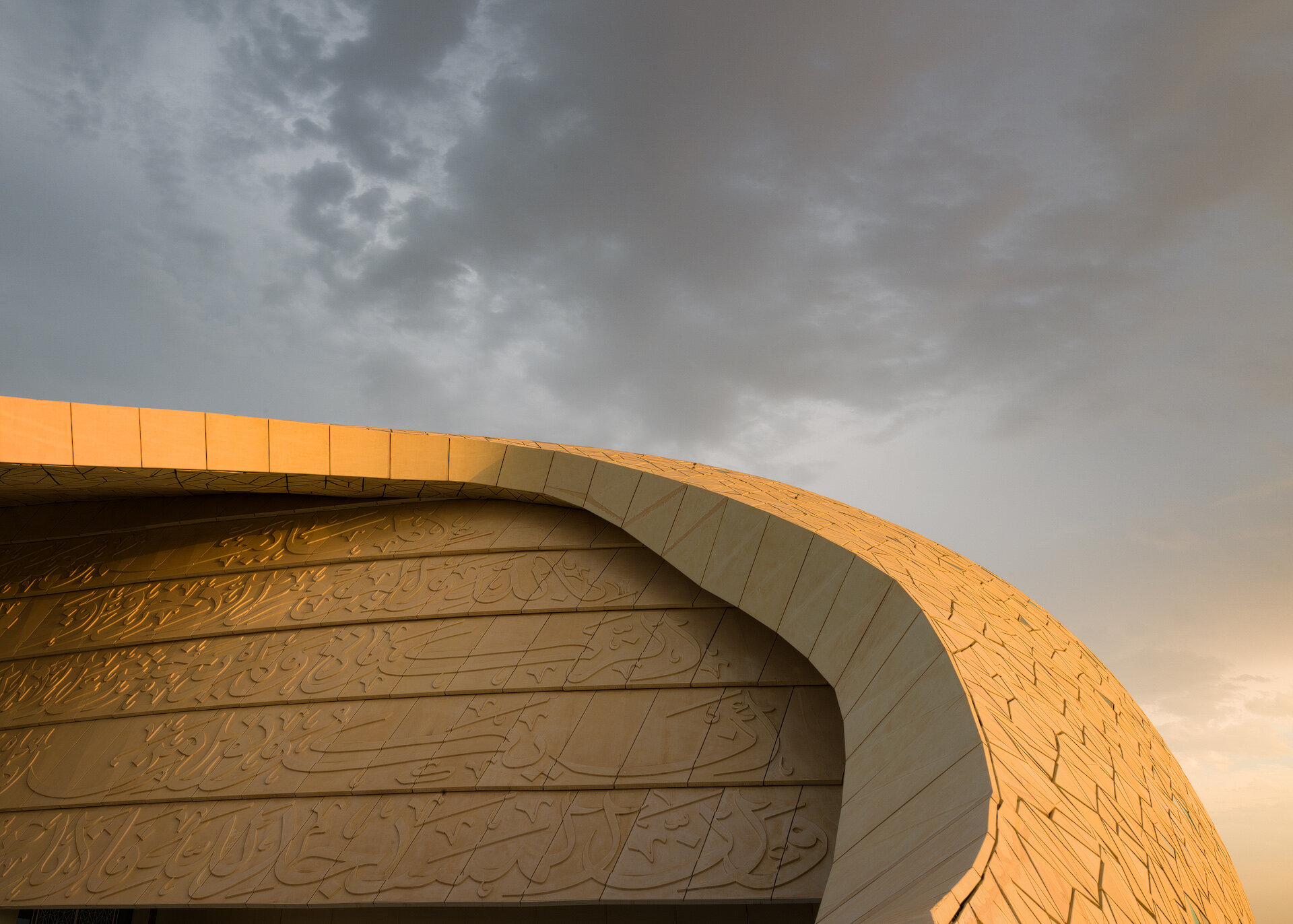 Detailed architectural shot of the Minaretein Center featuring embossed Arabic calligraphy on the curved wall lit by golden hour sunlight against overcast clouds.