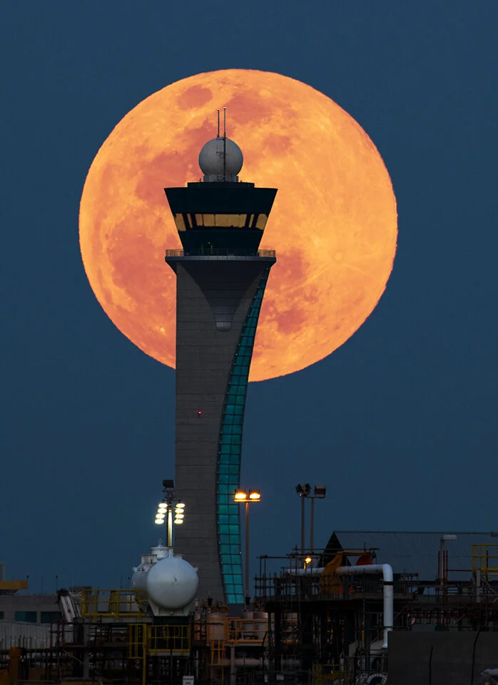 Telephoto photograph of a massive orange supermoon rising directly behind the Hamad International Airport control tower by Hammad Iqbal