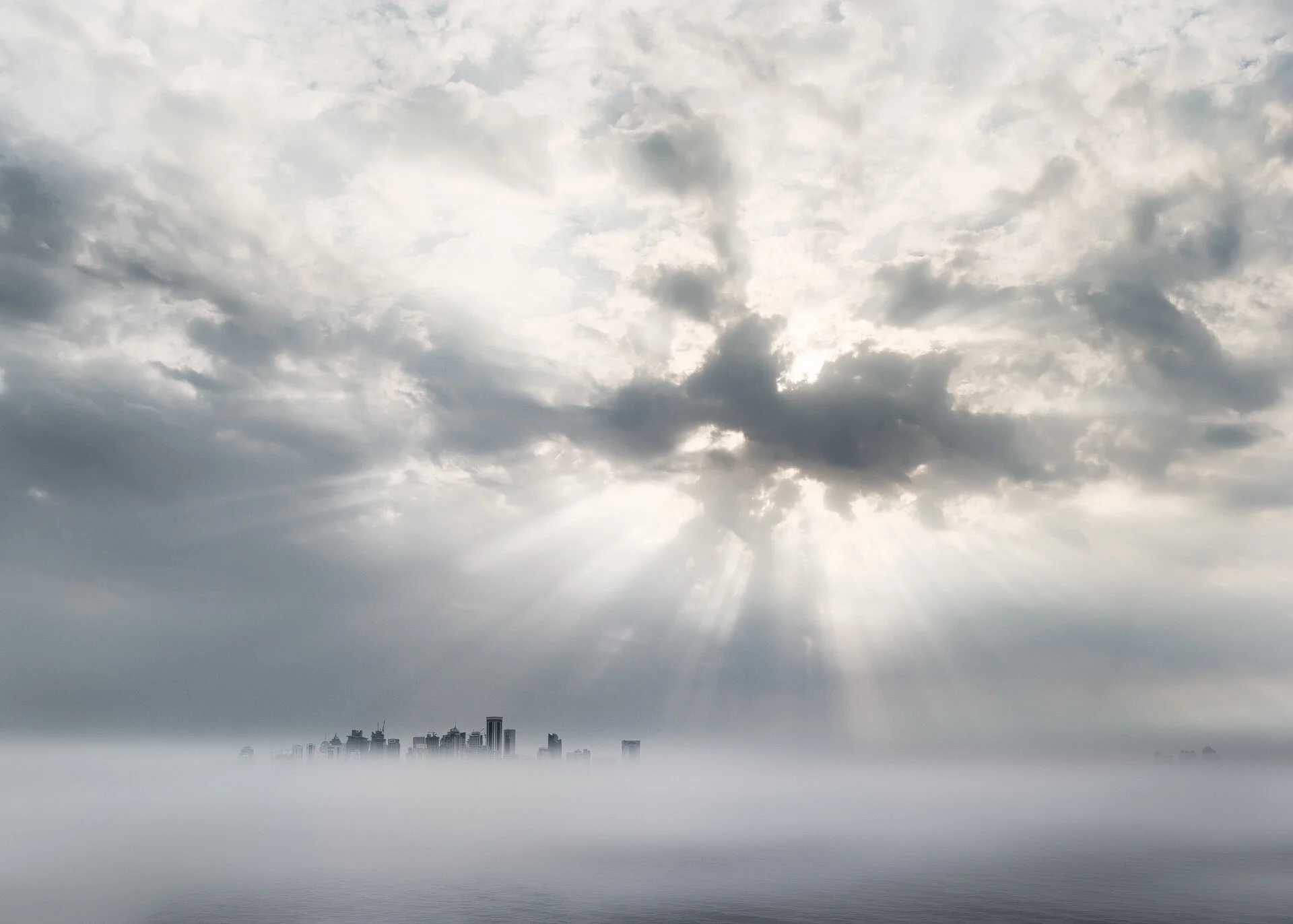 Photo of the Doha skyline against a dramatic cloudy sky, rays of sunlight radiating through clouds over a foggy city skyline.