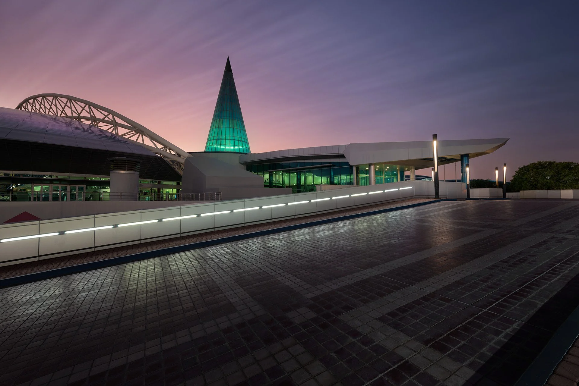 Architectural wide shot of the illuminated entrance ramp and glass facade of Aspire Dome showing the steel roof arch structure and green glass spire against a purple dusk sky.