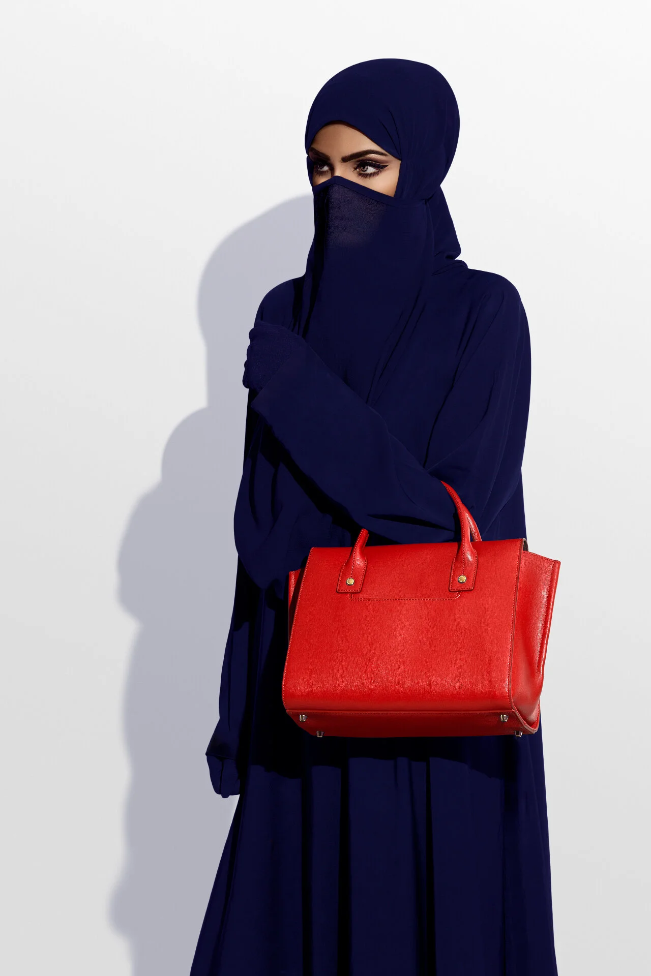 Studio fashion portrait of model holding a structured red leather handbag against navy abaya by Hammad Iqbal