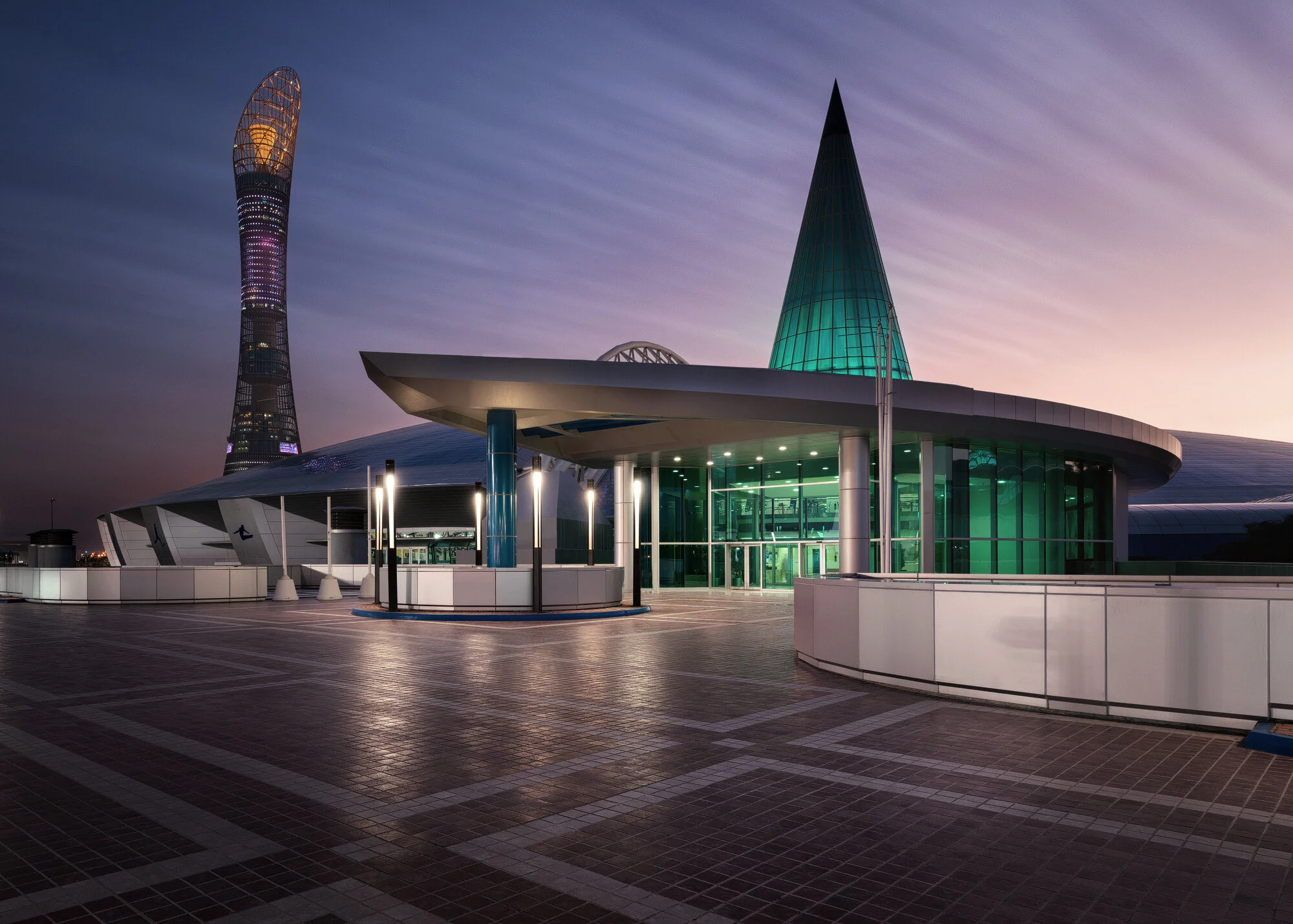 Exterior architectural photograph of the illuminated Aspire Dome entrance with green conical glass roof feature at twilight, with the 300-meter Aspire Tower standing in the background in Doha Sports City, Qatar.