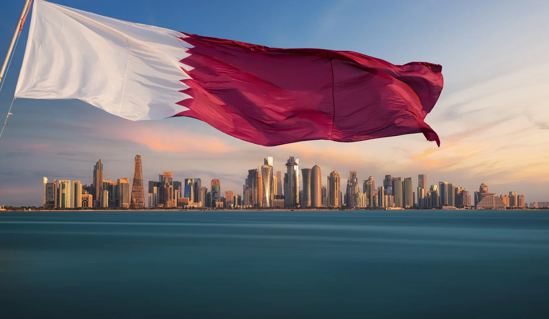 Qatari flag waving over calm waters with the illuminated West Bay skyline at sunset in Doha photographed by Hammad Iqbal