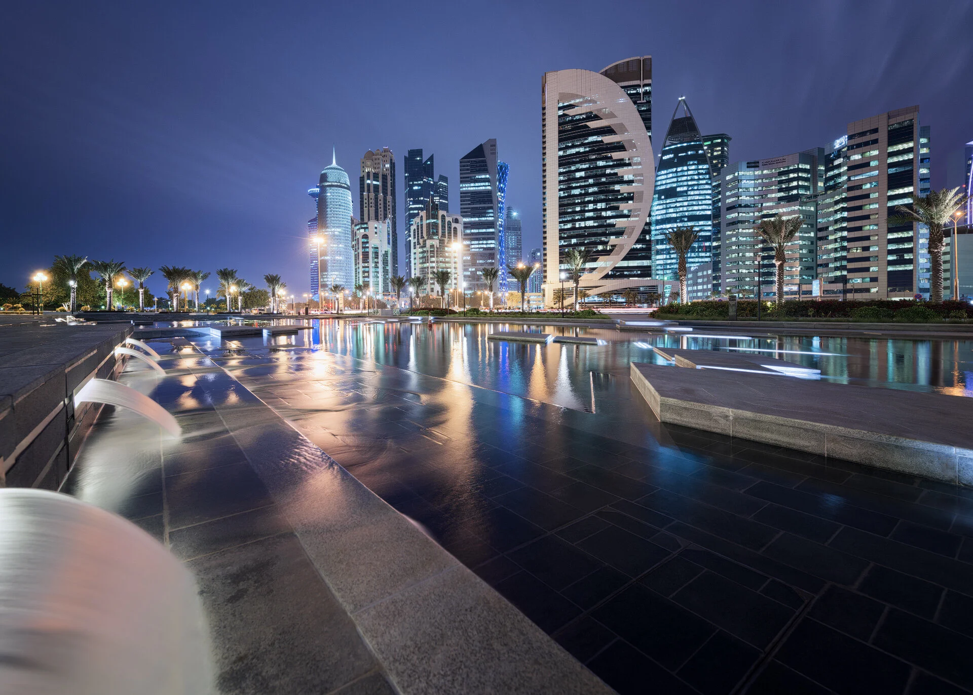 Wide night photo showing illuminated West Bay towers reflected across the calm water of the Hotel Park wading pool by Hammad Iqbal