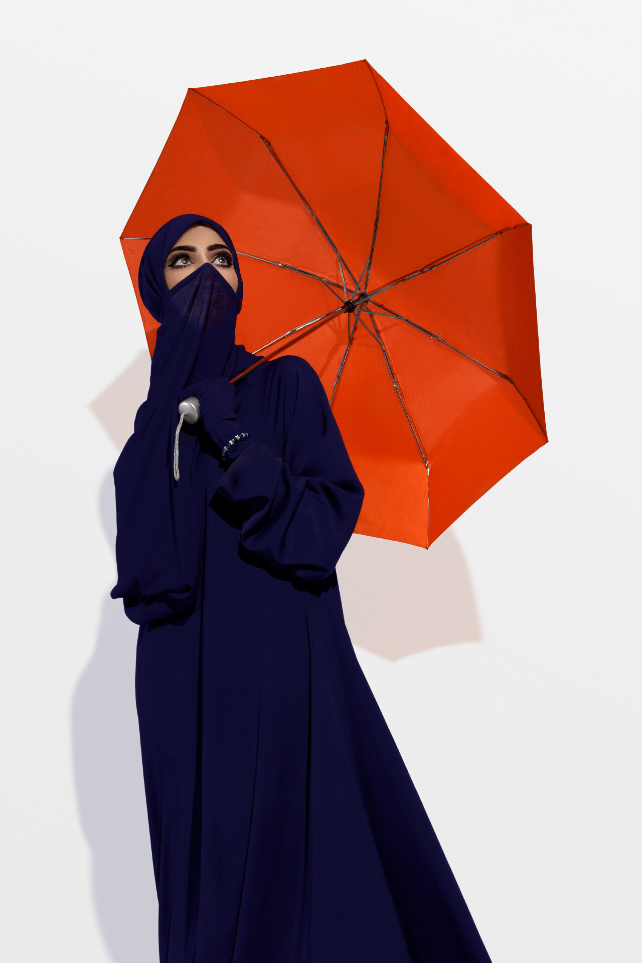 Three quarter studio portrait of modest fashion model with orange umbrella and crisp shadow against white wall by Hammad Iqbal