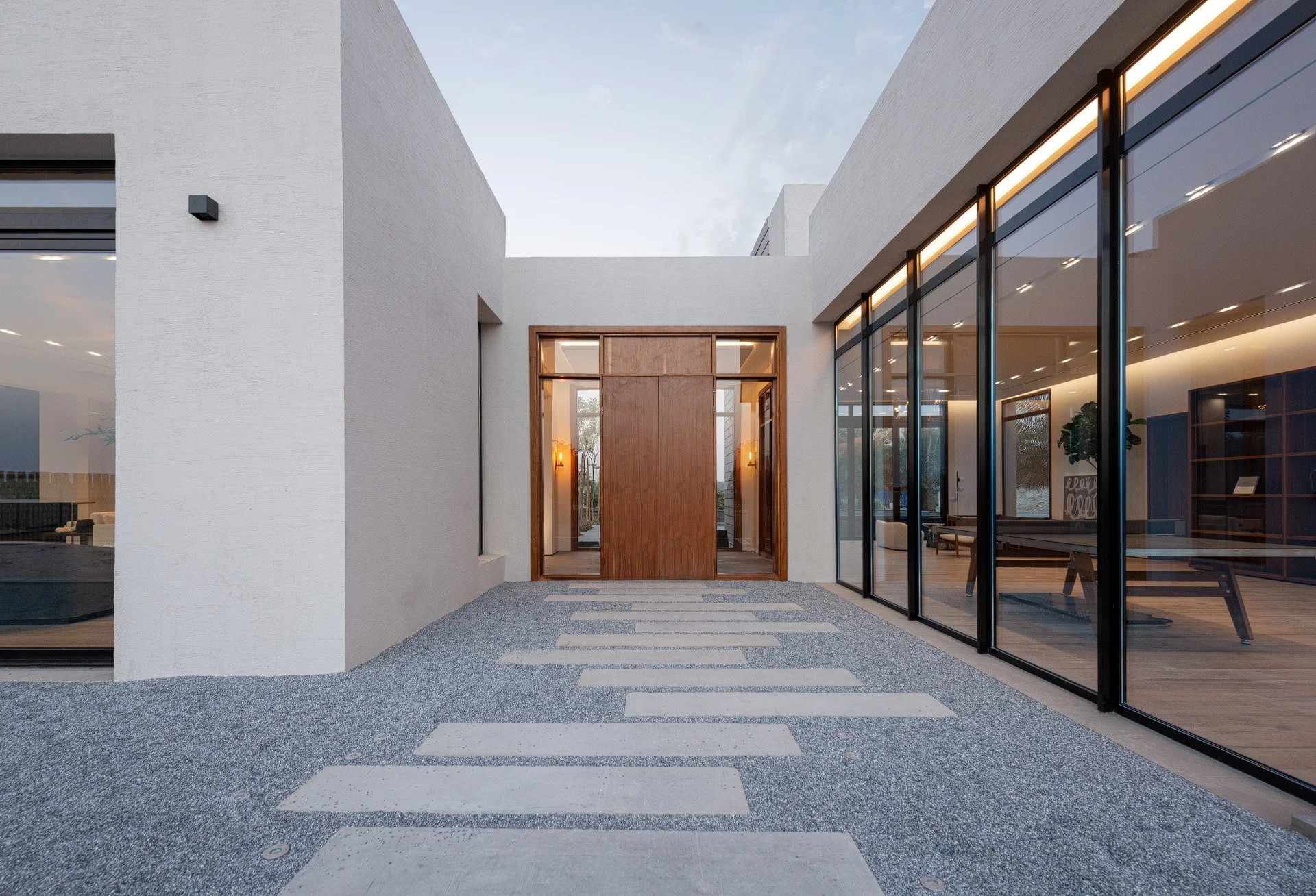 Textured gravel courtyard and floor-to-ceiling glass wall leading to a warm wood entrance door in a Qatar villa by Hammad Iqbal
