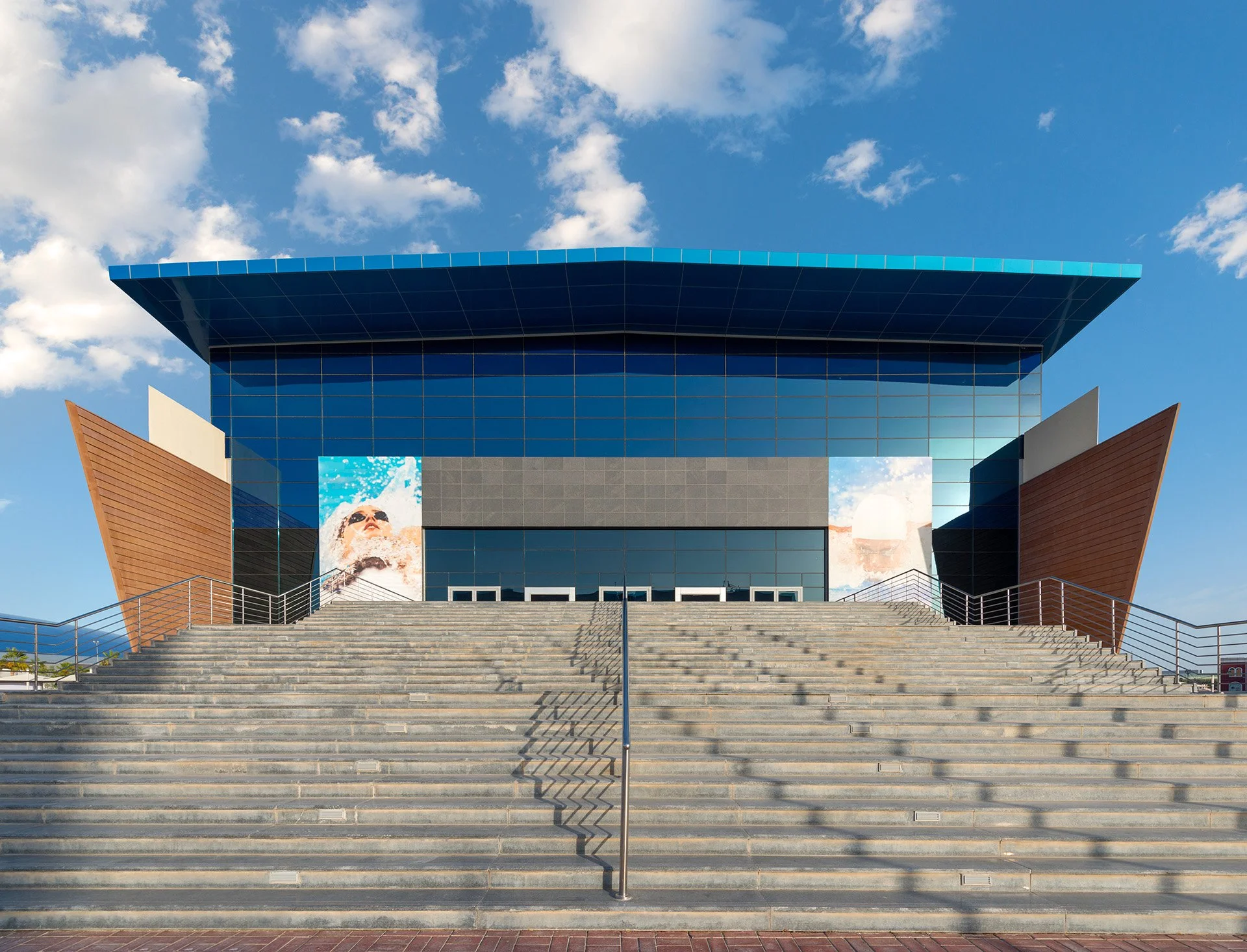 Symmetrical exterior architectural photograph of the entrance steps leading to Hamad Aquatic Center in Aspire Zone, featuring a blue glass facade and dynamic wing-like roofline under a blue sky.